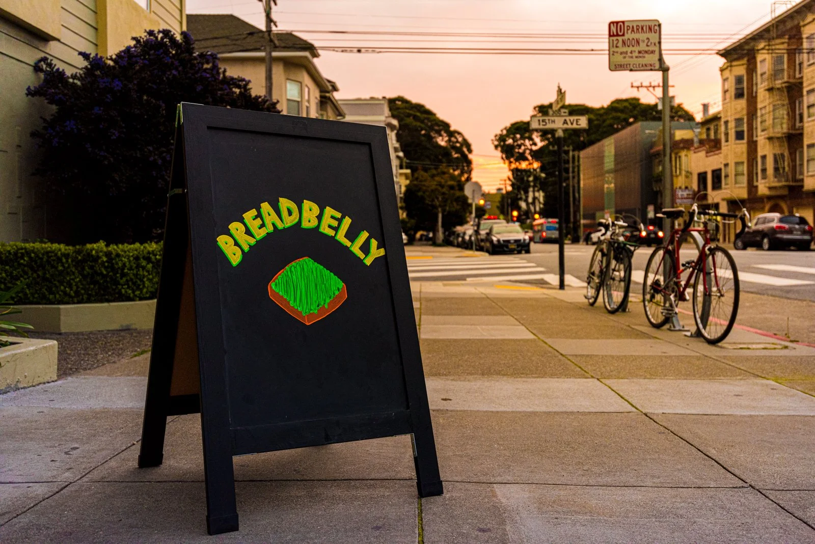 A sidewalk sidewalk sign advertising Breadbelly with a drawing of a bread loaf topped with green spread, on a city street at sunset.