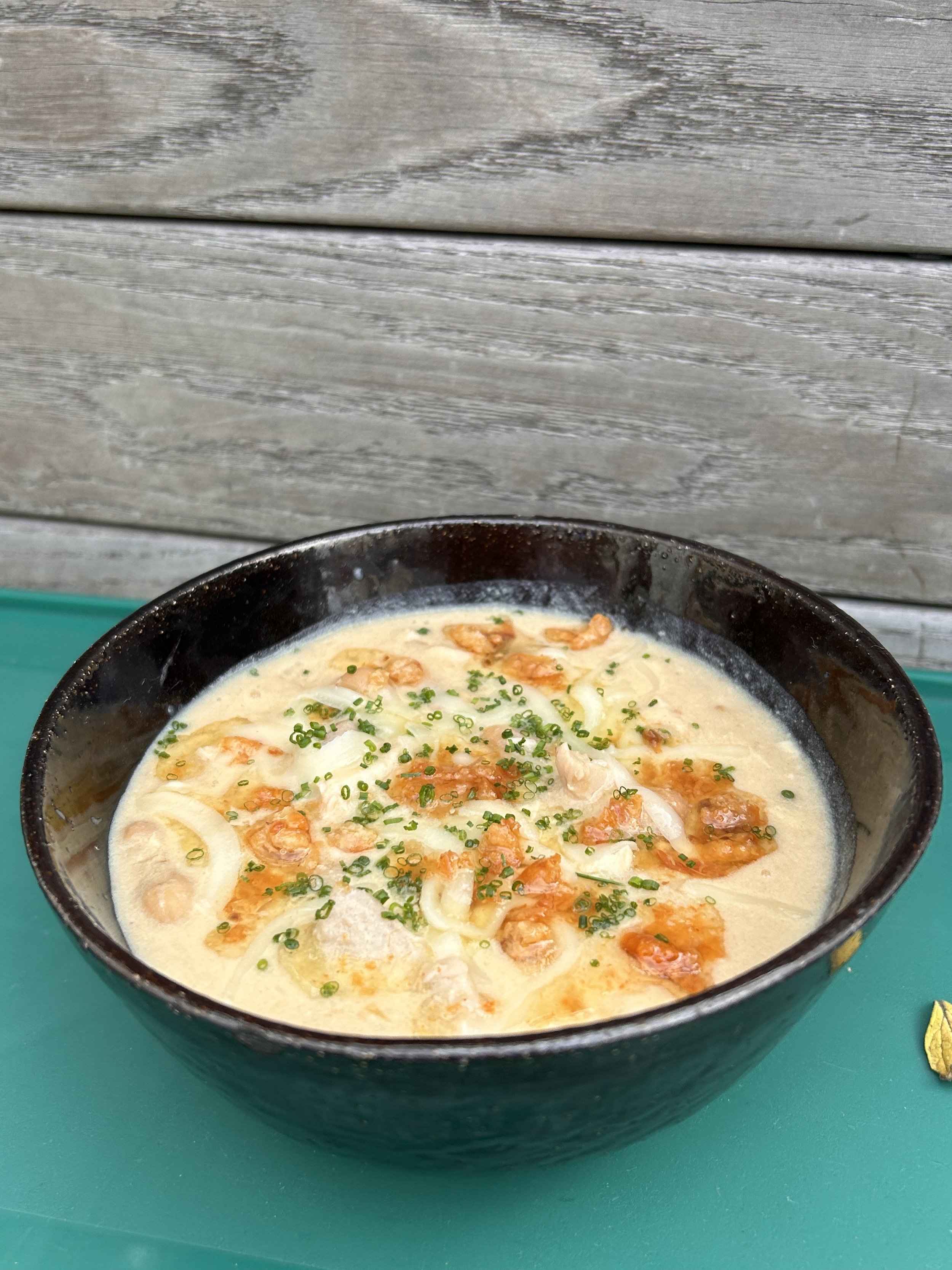Bowl of creamy seafood soup garnished with chopped chives, served in a black bowl on a teal surface, with a weathered wooden backdrop.