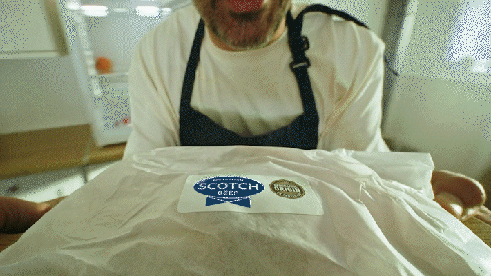 Man wearing a white shirt and black apron holding a wrapped package labeled 'Scotch Beef' and 'Origin' in a kitchen.