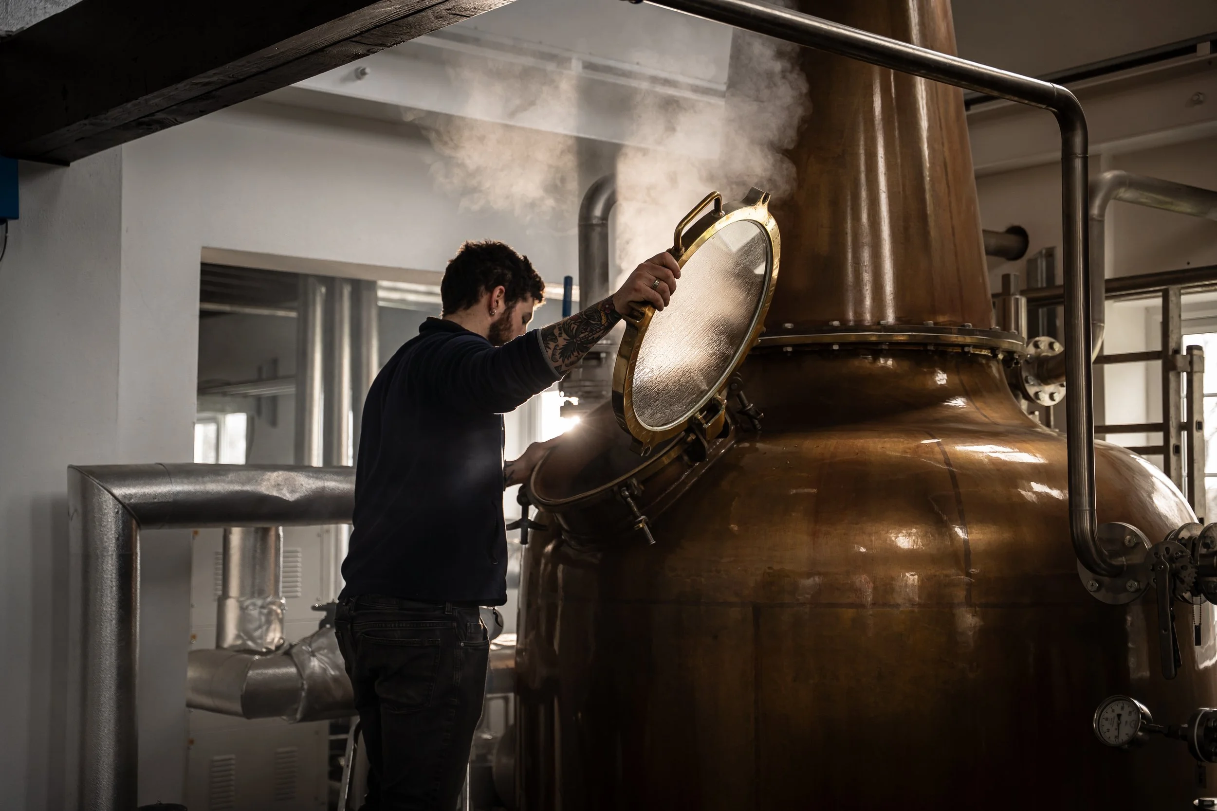 Man with tattoos opening a large metallic lid on a copper still in a brewery or distillery, with visible pipes and steam.