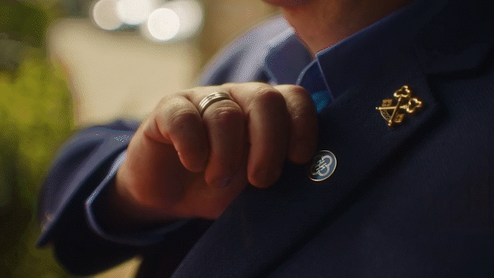 Close-up of a person adjusting their blue suit jacket, showing a gold pin and a lapel button.