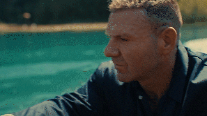 A man with short blonde hair and a serious expression looking down, dressed in a dark blue shirt, outdoors near water with trees in the background.