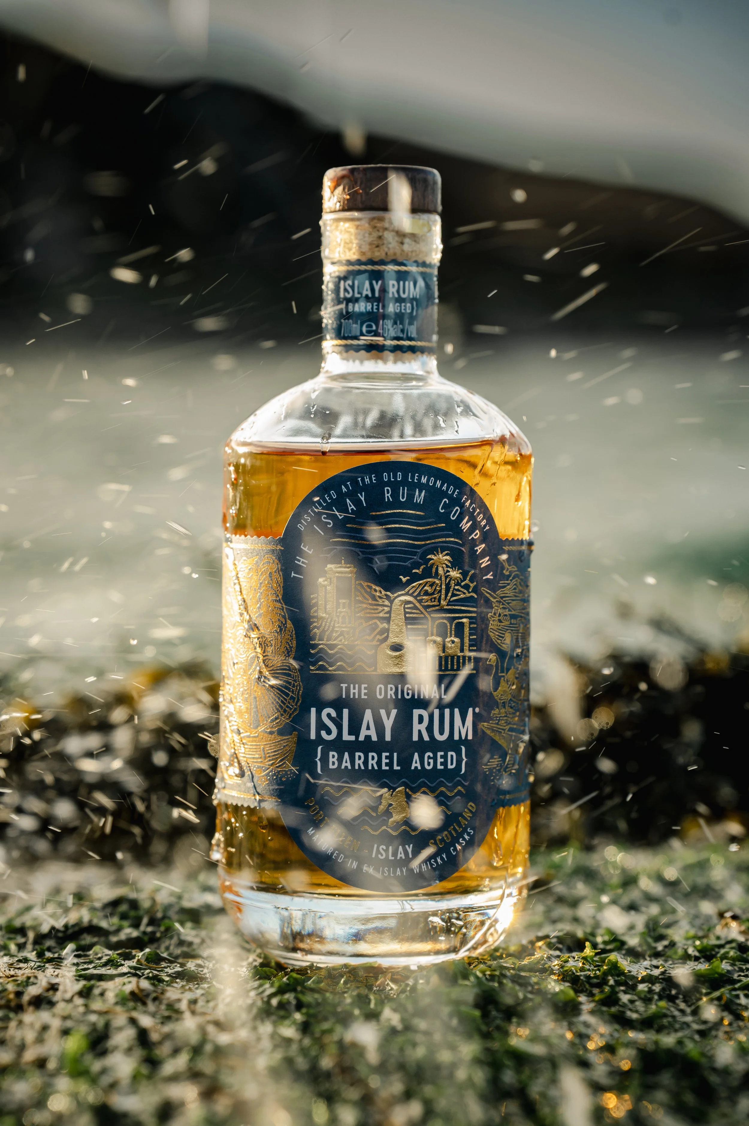 A bottle of Isle of Rum barrel-aged rum standing on the ground outdoors, with water splashing around and sunlight shining on it.