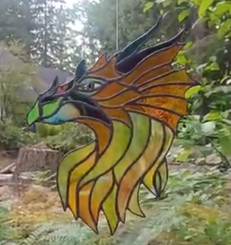 Colorful stained glass art of a dragon's head with a green eye and orange and yellow mane, hanging outdoors among green leaves.