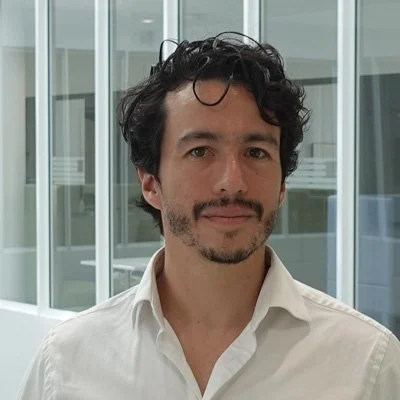 A headshot image of David R. Goyes with short brown hair, close-cropped beard and moustache, wearing an open-necked white shirt..
