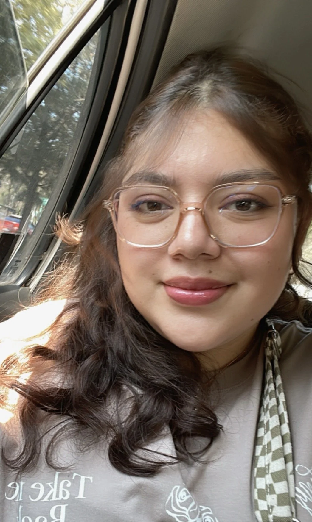 A woman with long, wavy dark hair wearing glasses, smiling, sitting by a window on a bus during daytime.