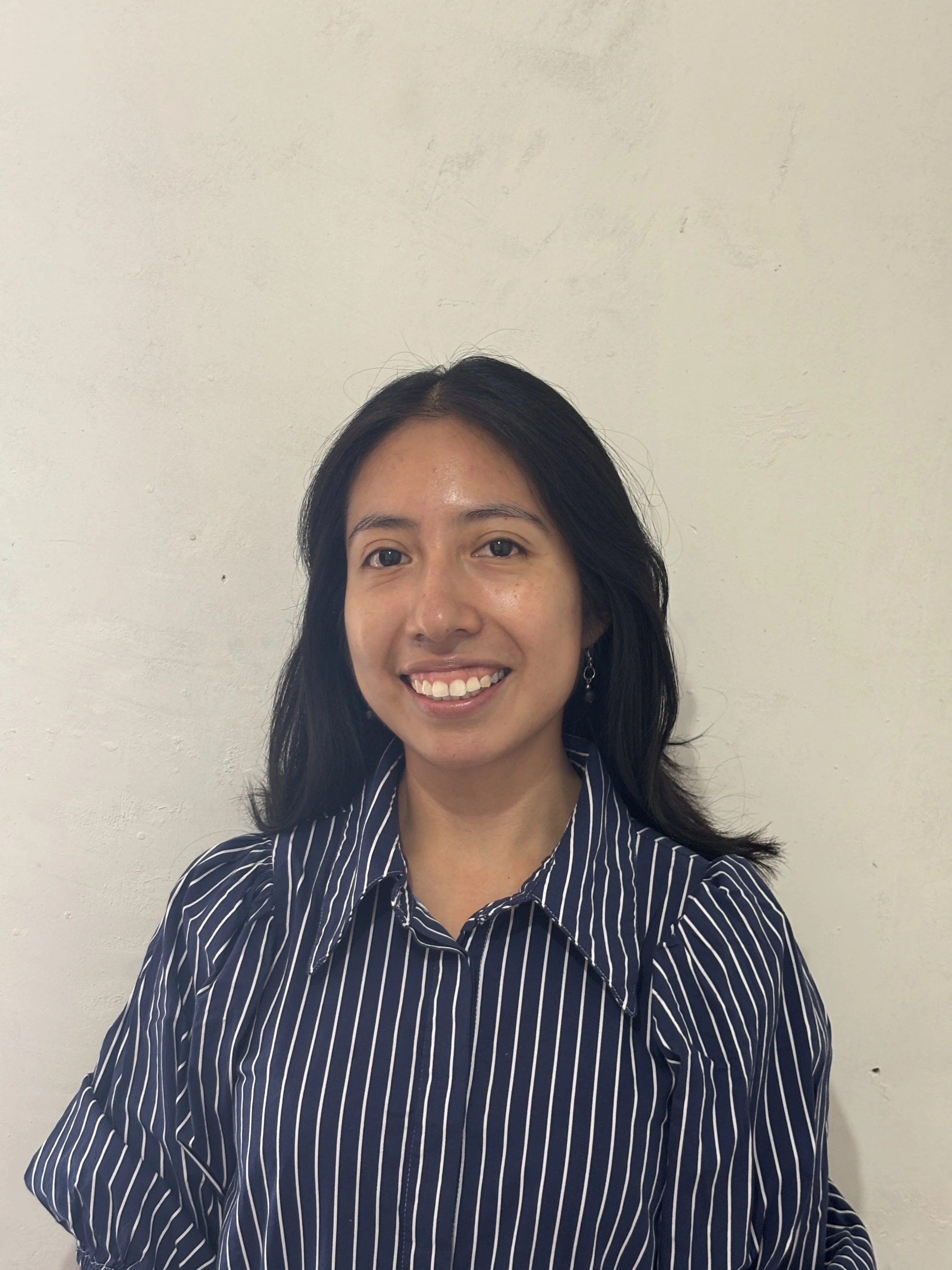 A young woman with long dark hair smiling and hugging a tree in a sunny, wooded park with green grass and tall trees.