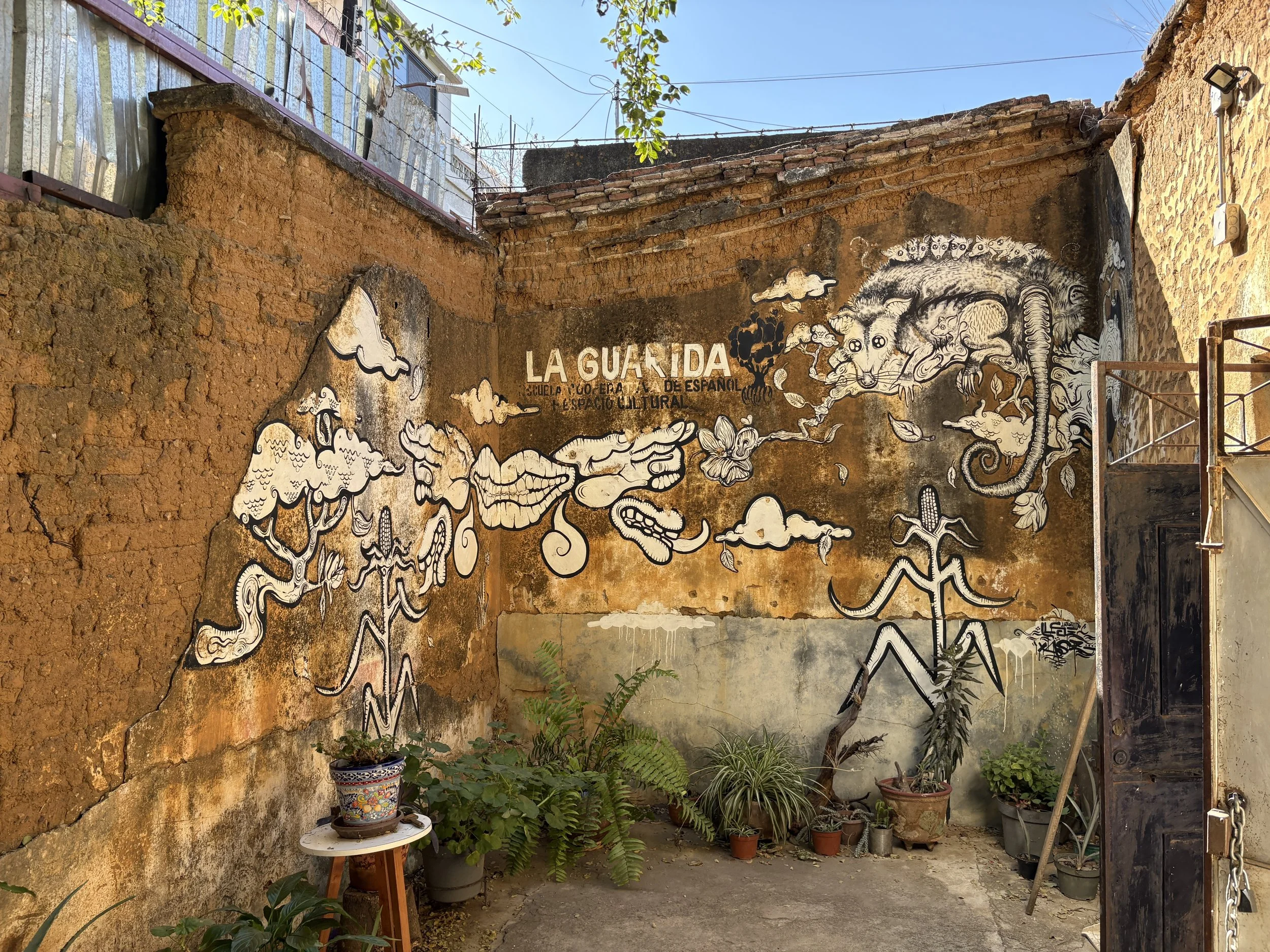 Mural on a weathered brick wall featuring cartoonish illustrations of people, animals, and clouds, with the words "LA GUARDIA" and Spanish text underneath, in an outdoor courtyard with potted plants.
