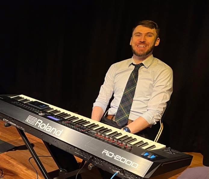 Richard sitting at a digital piano on the stage. He is in his 30s, has brown hair, a short brown beard, a tartan tie and is smiling.