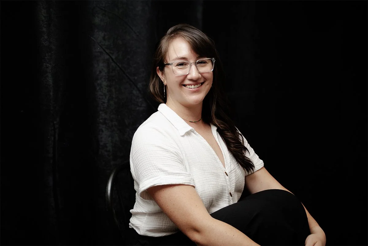 black backdrop, a woman designer sitting upright wearing glasses, her hair down and a white blouse