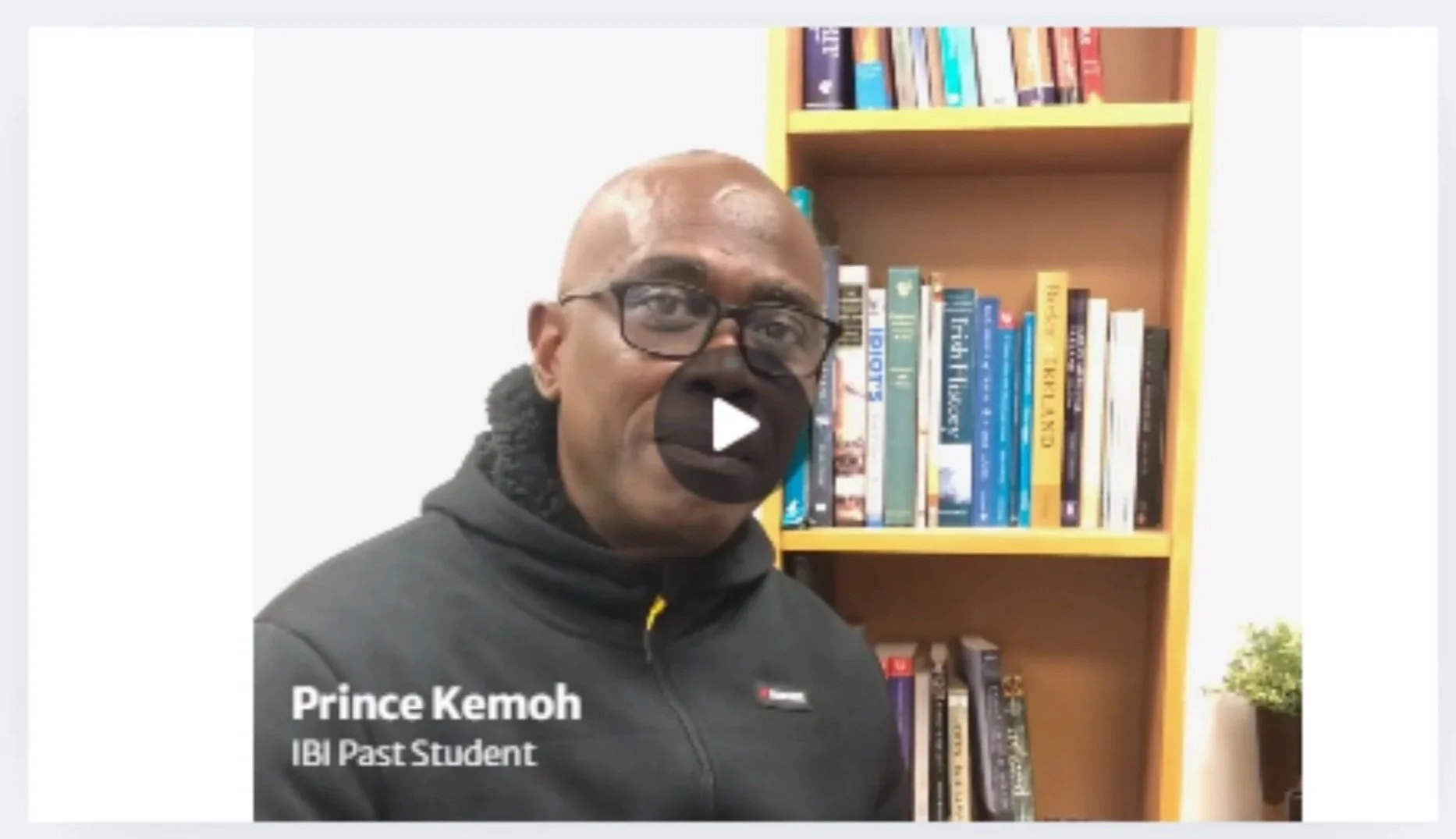 A man with glasses and a black hoodie standing in front of a bookshelf filled with books and a small potted plant on the side. The text overlay says 'Prince Kemoh, IBI Past Student'.