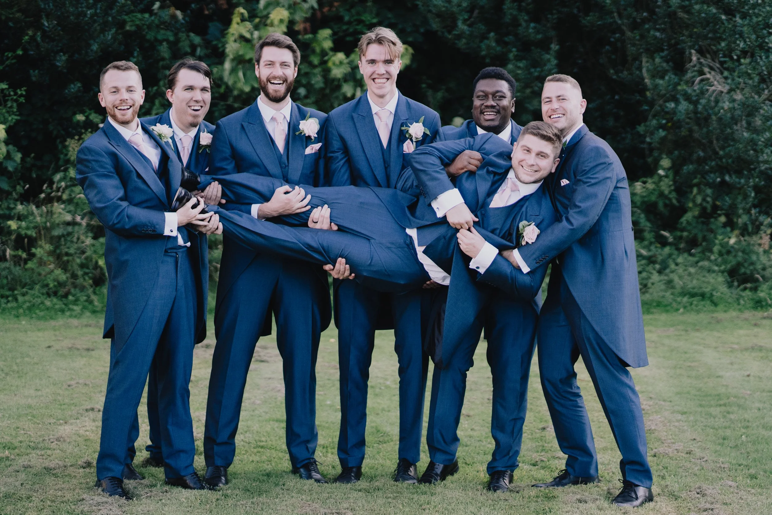 Eight men dressed in blue suits outdoors, holding one man horizontally in the center, all smiling.
