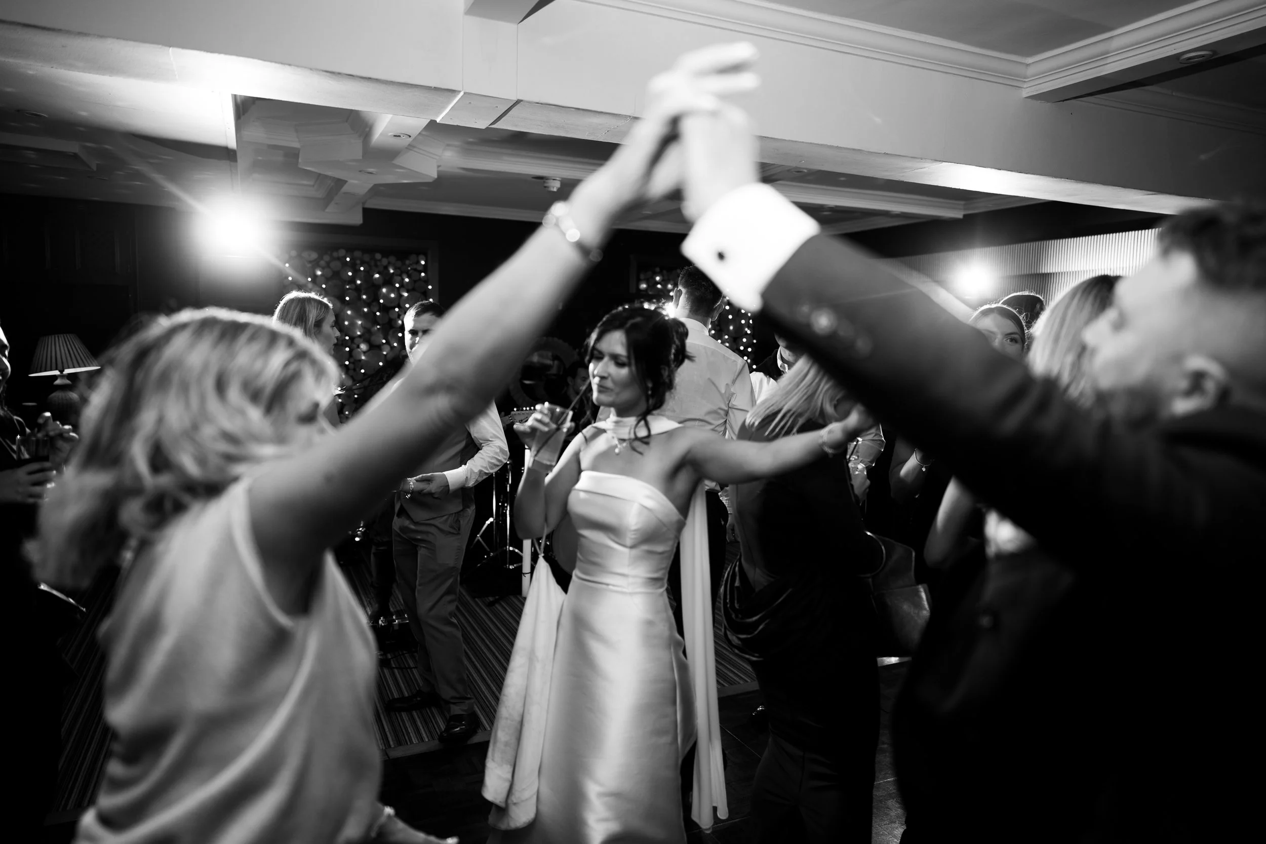 A woman in a strapless dress drinks from a straw while dancing at a crowded party with people dancing and socializing in a dimly lit room with string lights and wall decorations.
