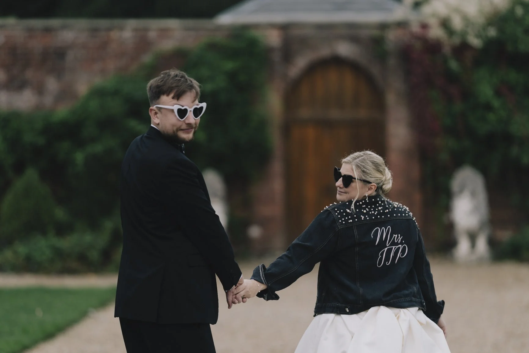 A couple holding hands outdoors, dressed in wedding or formal attire, with the woman wearing a denim jacket with "Mrs. Trp" embroidered on the back and sunglasses, and the man in a black suit and heart-shaped sunglasses.