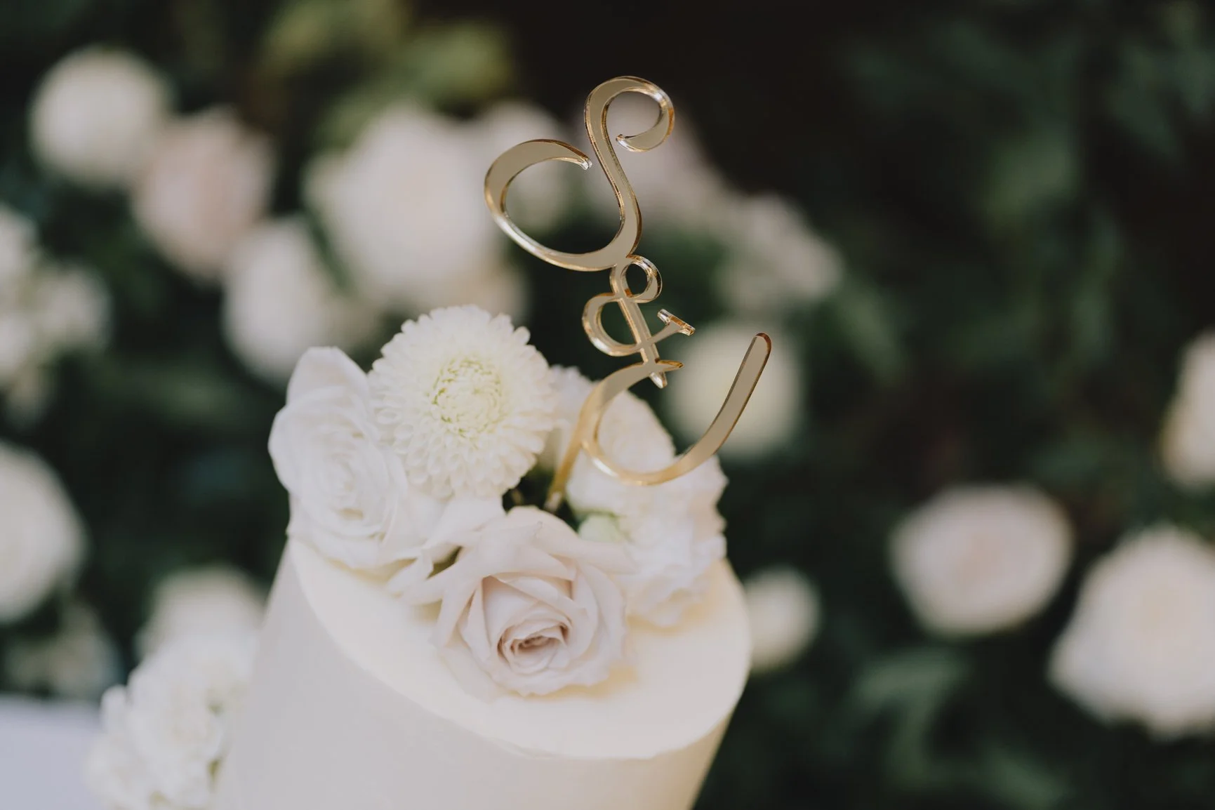 A white wedding cake decorated with white roses, a white chrysanthemum, and a gold "Love" cake topper, with a blurred background of greenery and white flowers by Blooms at Breach