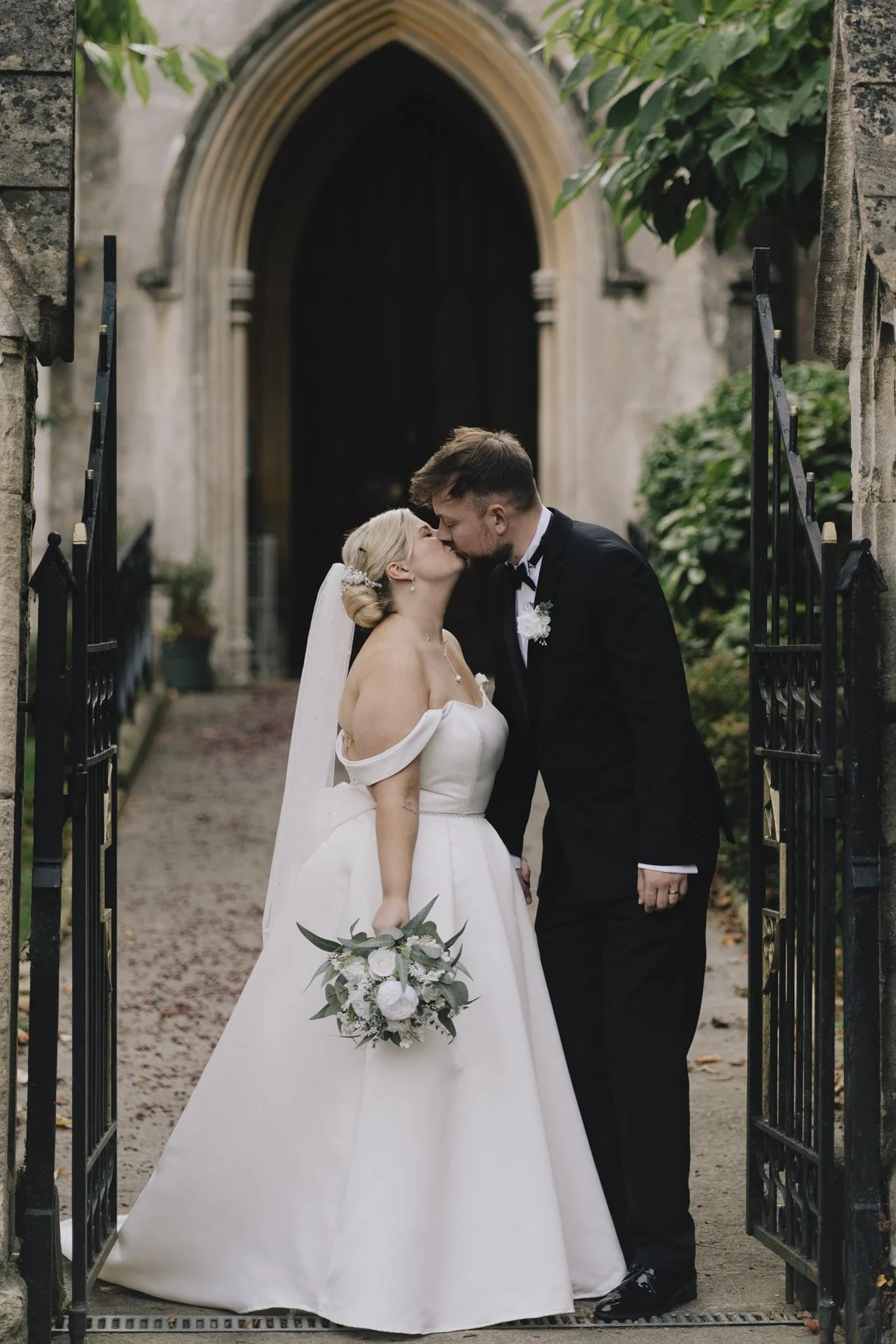 A bride and groom share a kiss outside a stone church with an arched doorway, surrounded by greenery taken by Paulton Wedding Photography