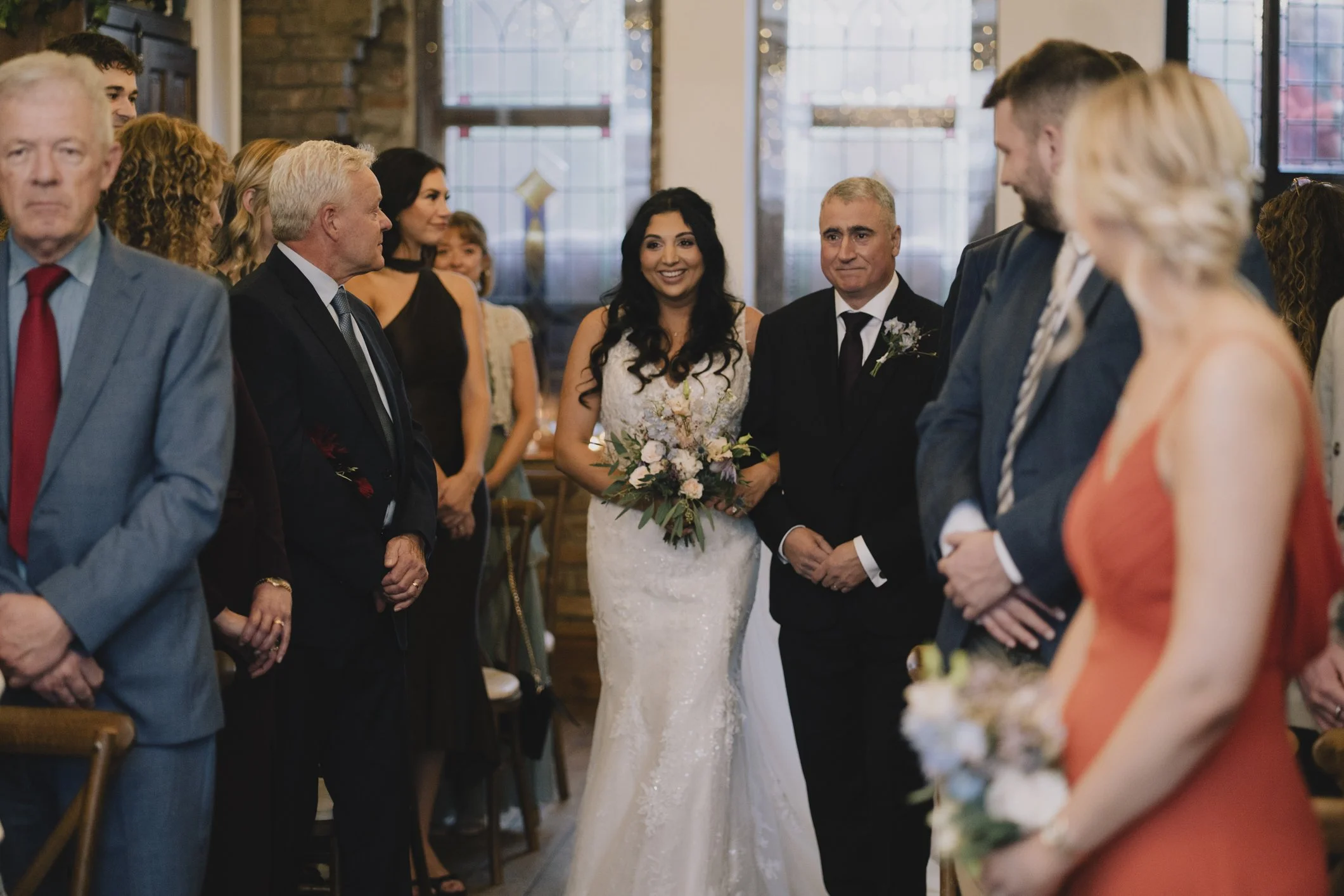 A wedding ceremony with a bride in a white lace gown holding a bouquet, standing next to a groom in a dark suit. They are surrounded by seated and standing guests dressed in formal attire, inside a  Bristol venue with large windows and brick walls.