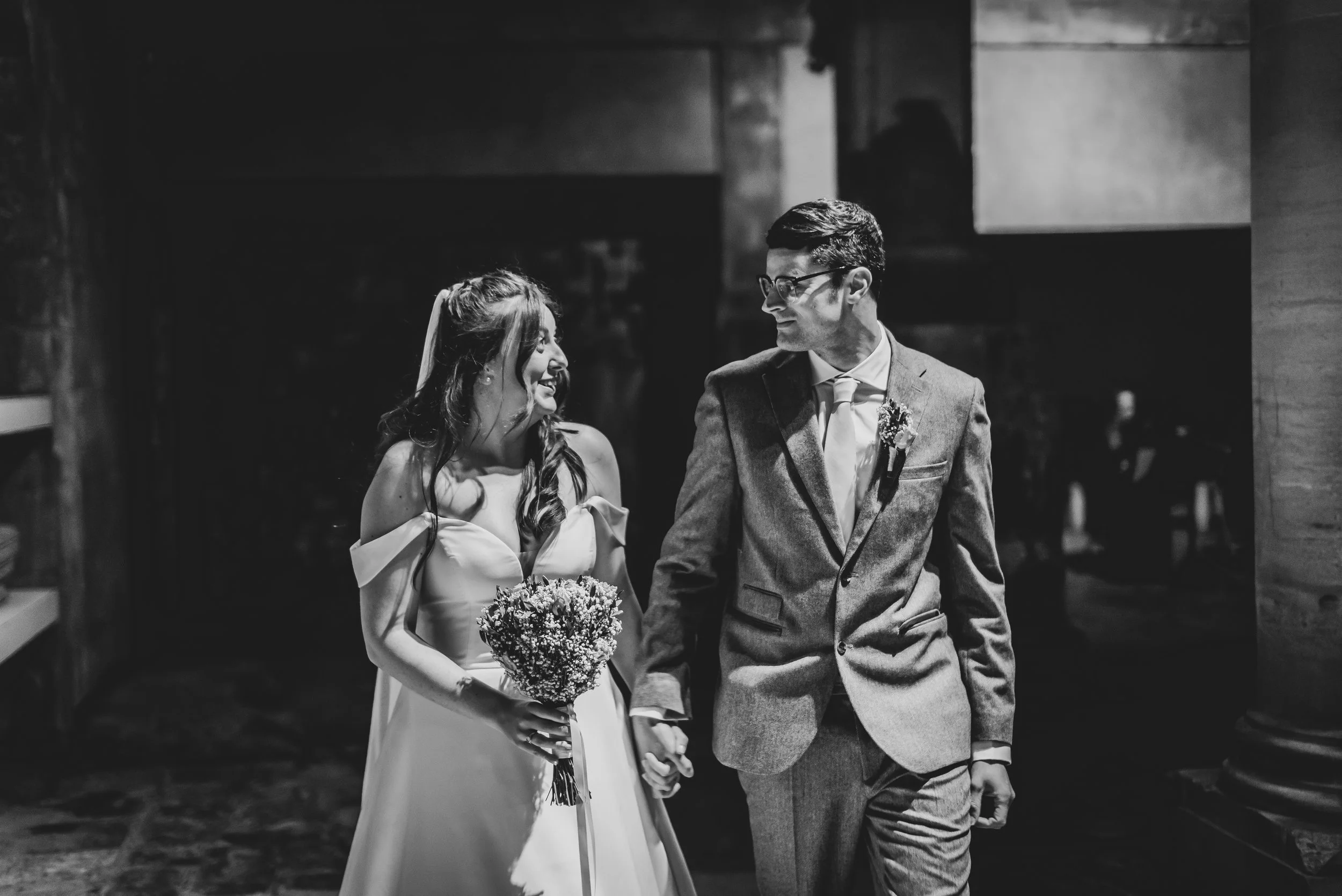 A black and white photo of a bride and groom holding hands, smiling at each other, walking inside a building at Roman Baths