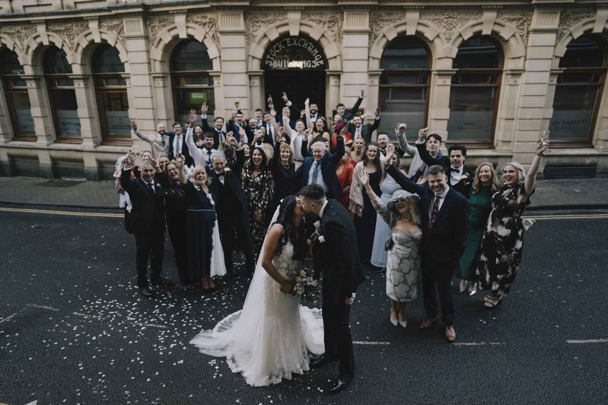 A wedding celebration with a bride and groom kissing in front of a crowd of guests outside a building.