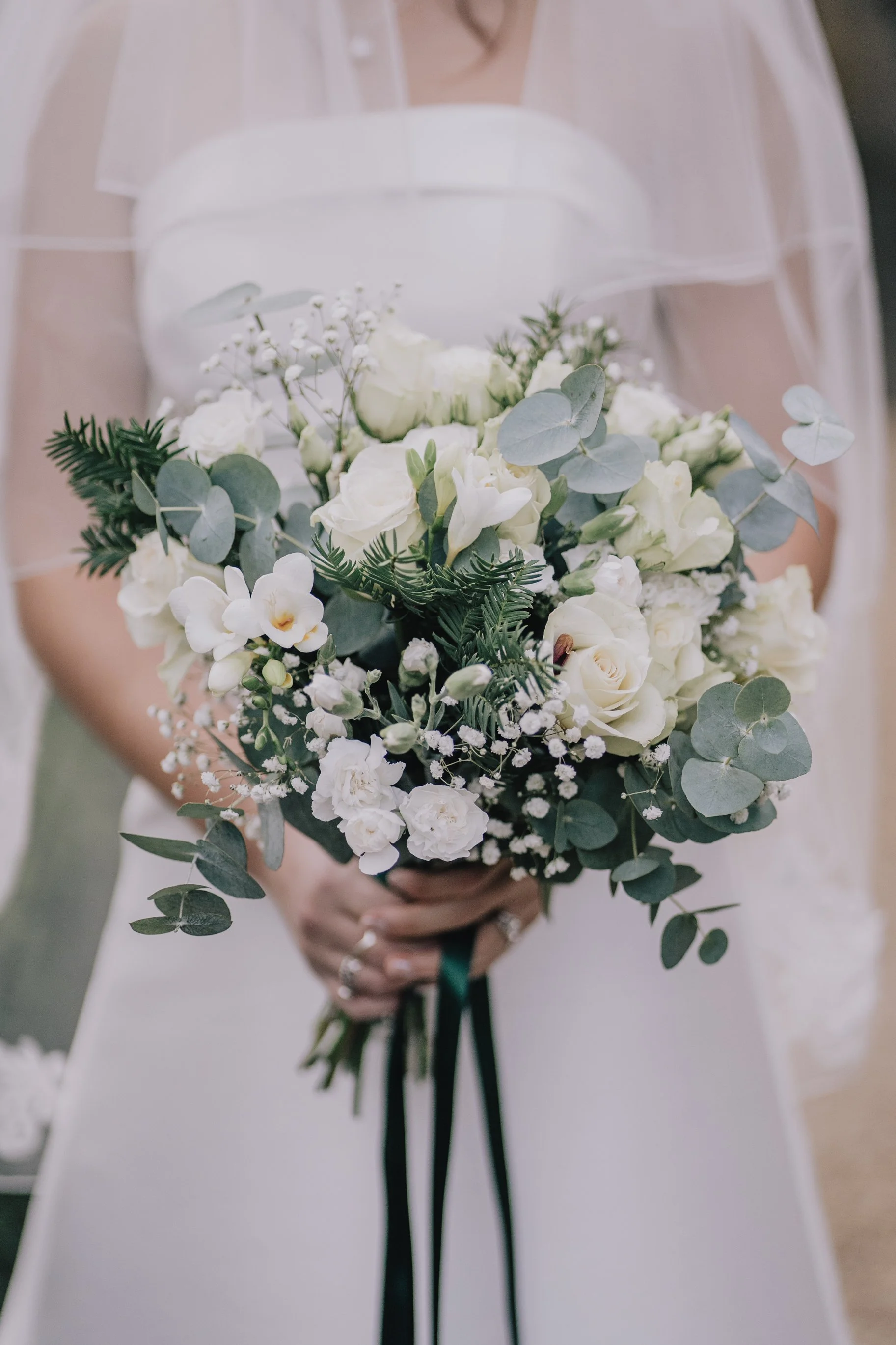 A bride in a white dress holding a bouquet of white flowers and greenery, with a veil draped over their shoulders