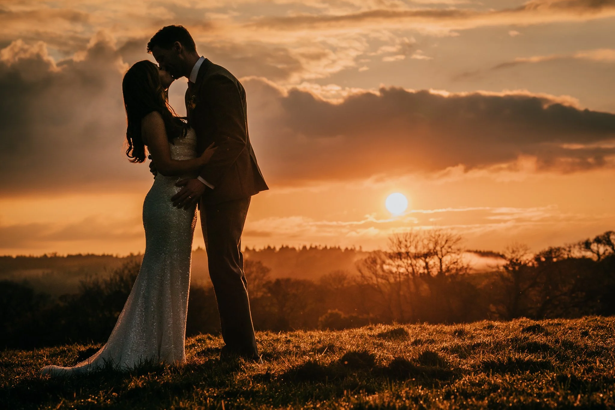 A couple in wedding attire embracing outdoors at sunset, with the sun low in the sky and clouds above, and trees in the background.