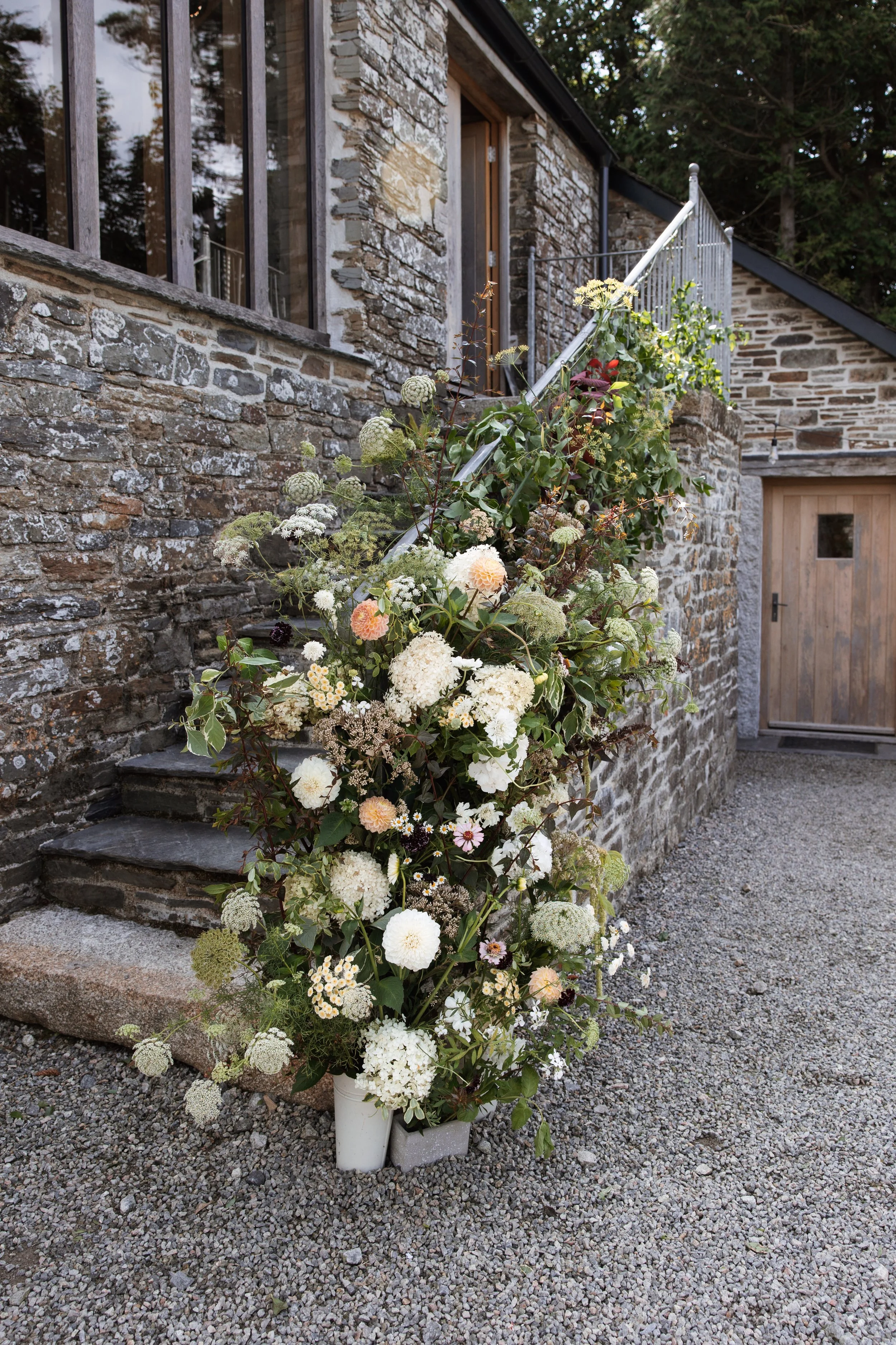 A large floral arrangement with white and pastel-colored flowers placed at the base of stone steps outside a rustic stone house with wooden window frames and a wooden door.