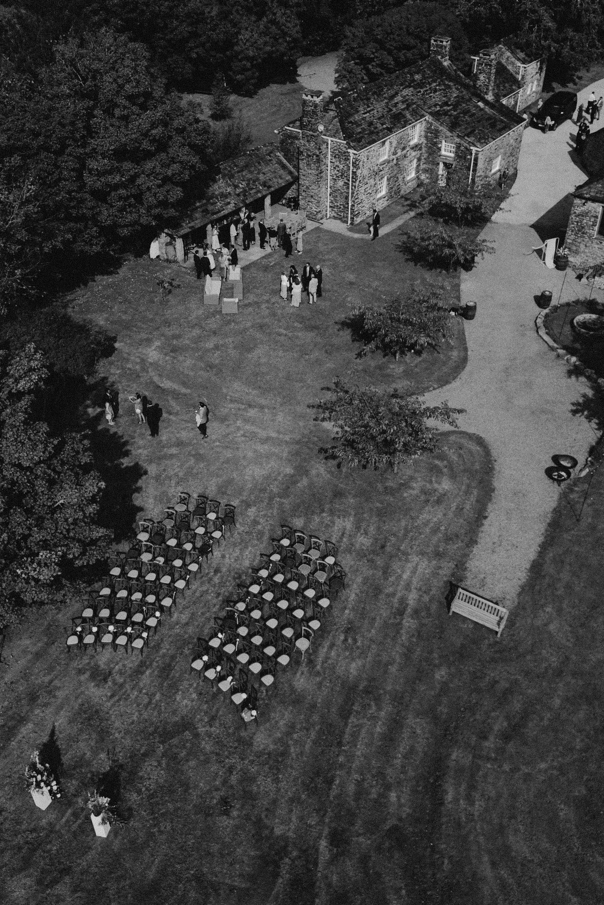 An outdoor wedding reception scene in black and white, with tables and chairs set up on grass, and a group of people gathered near a rustic stone building surrounded by trees.