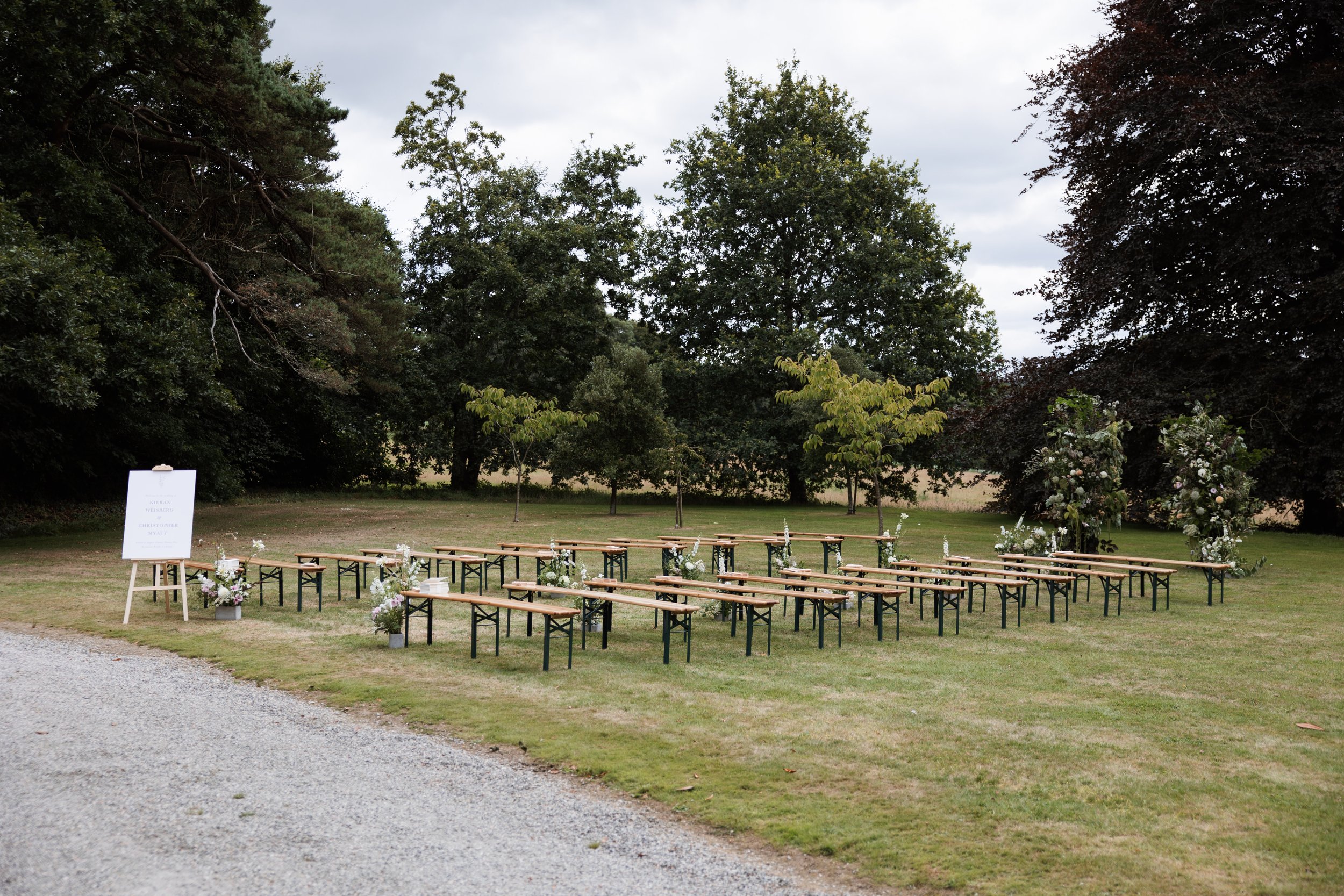 Outdoor wedding ceremony setup with benches arranged in rows on a grassy area, decorated with white flowers and greenery, with trees and a cloudy sky in the background.