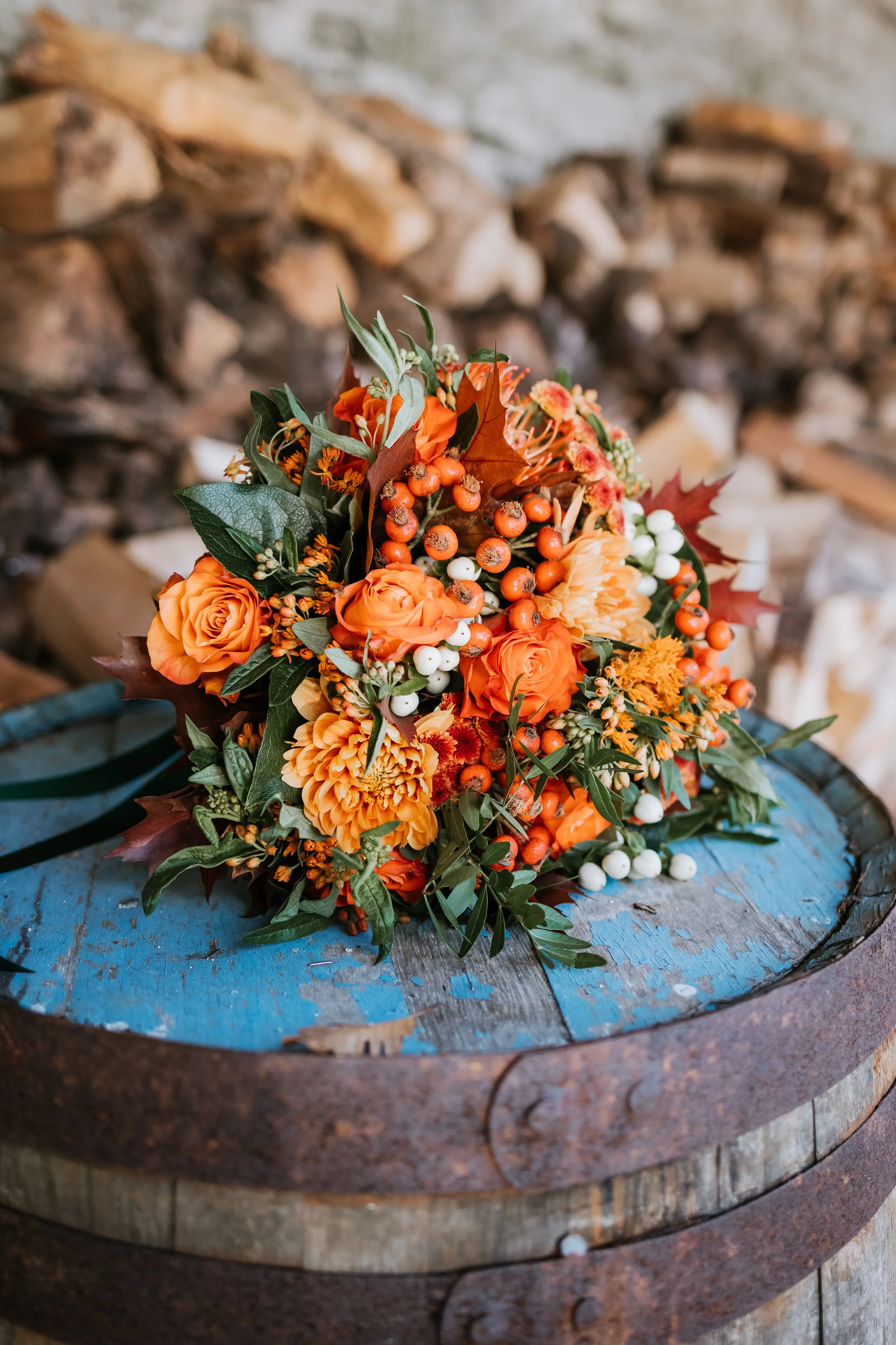 A vibrant orange and green floral bouquet with roses, dahlias, berries, and leaves rests on a weathered blue wooden barrel.