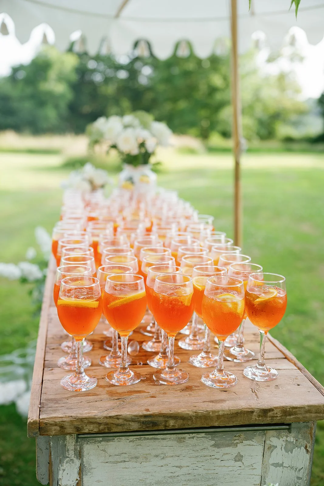 A row of glasses filled with orange-colored drinks with lemon slices on a rustic outdoor table at a summer event.