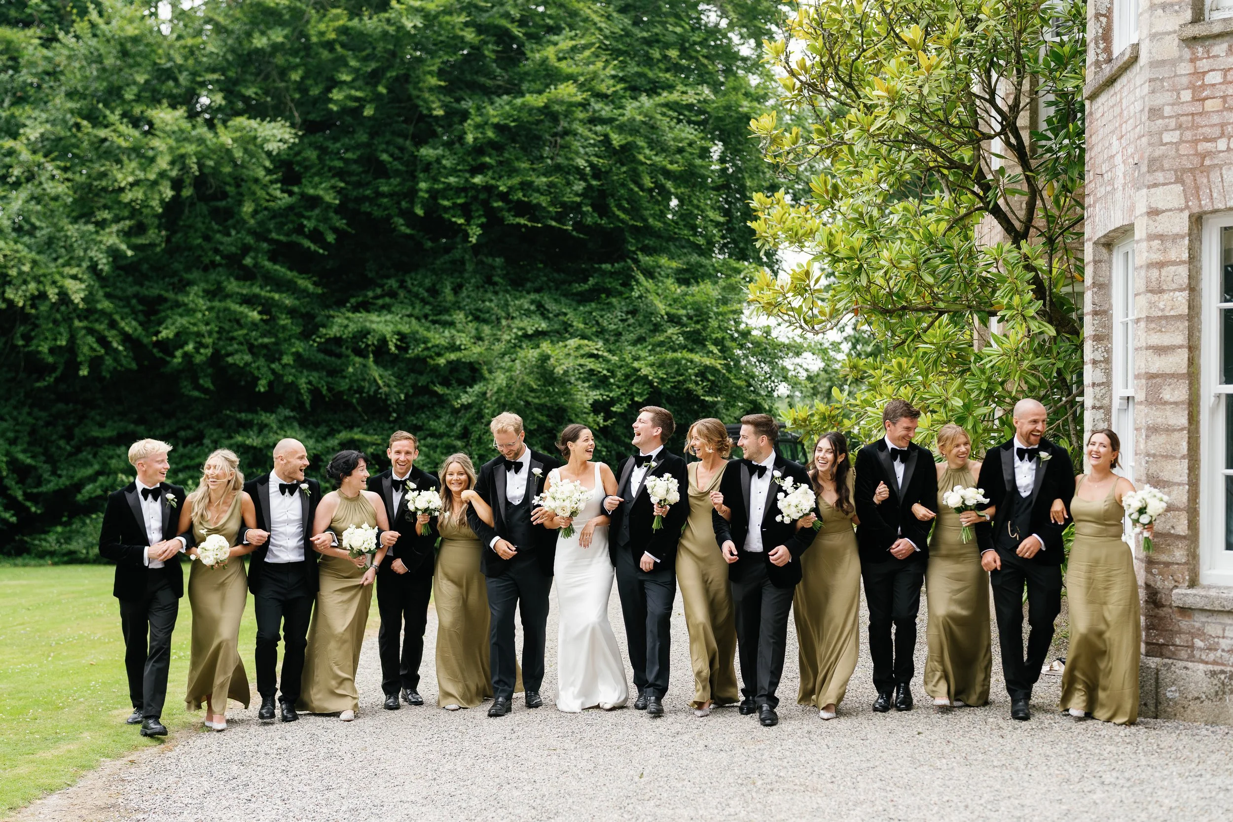 Wedding party of 14 people walking outdoors, with the bride and groom in the center, surrounded by bridesmaids and groomsmen, all smiling and laughing.