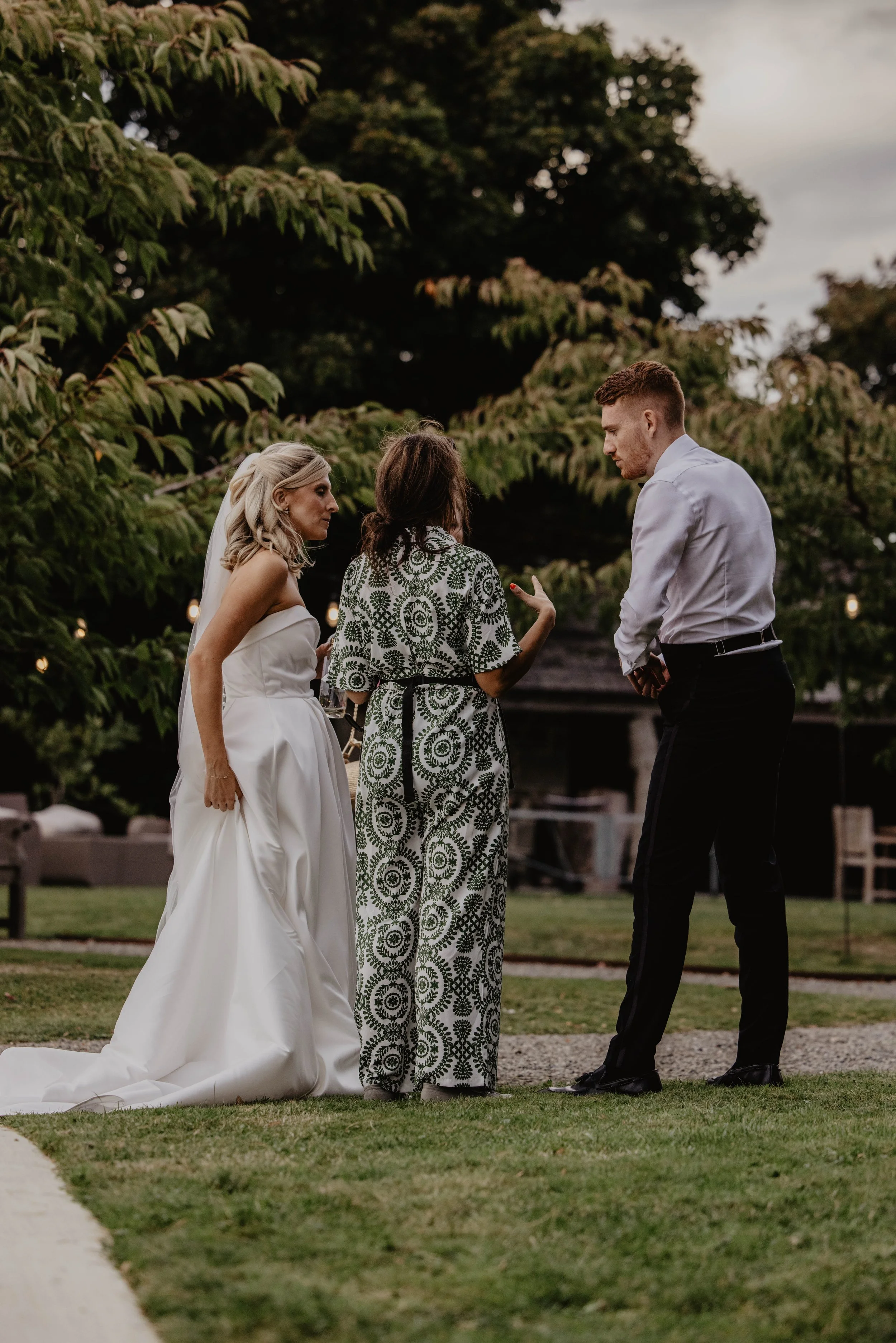 A bride, an officiant, and a groom standing outdoors during a wedding ceremony, with green trees and a cloudy sky in the background.