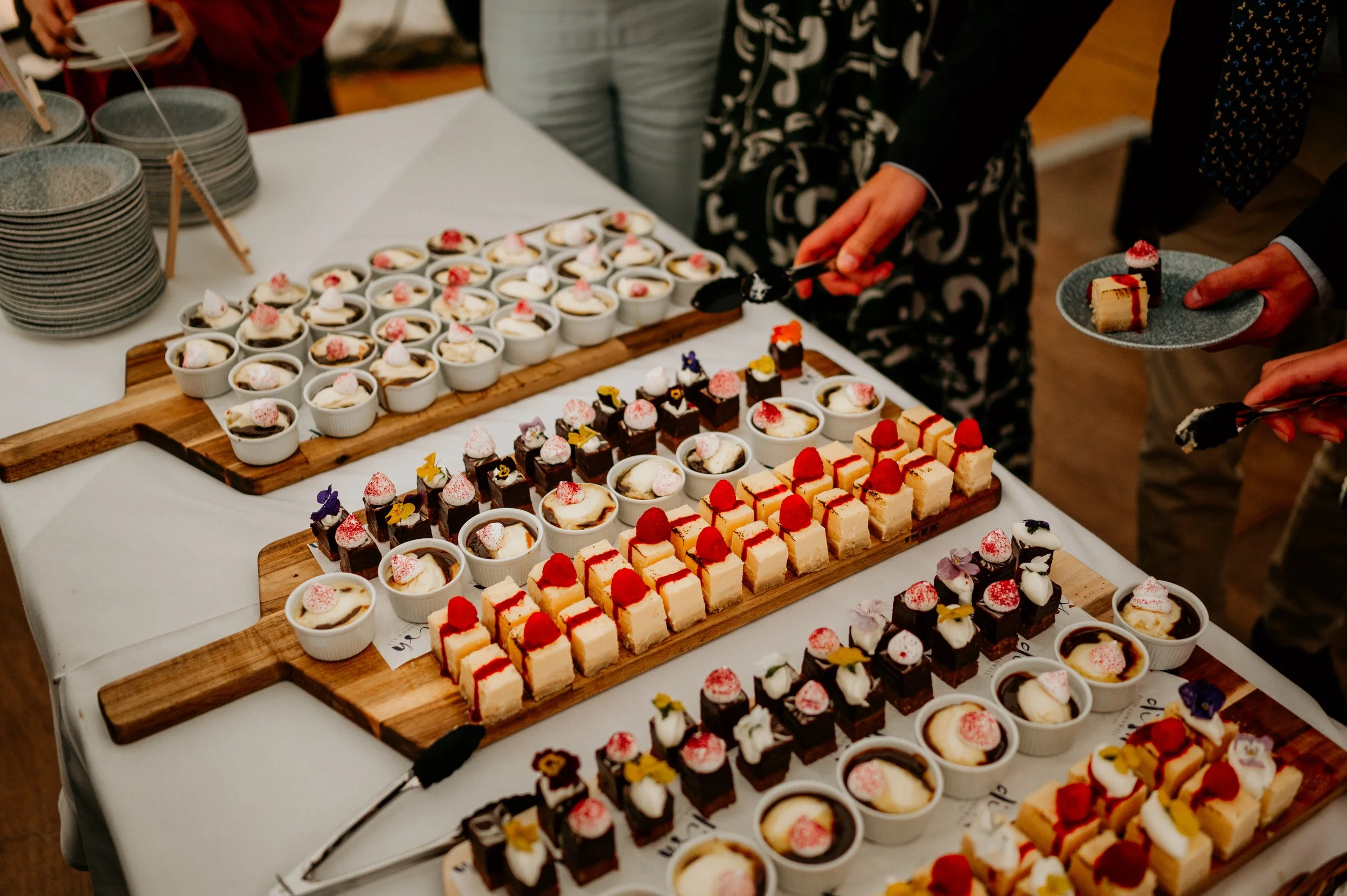 Assorted small desserts including cheesecake squares, mousse cups, and chocolate confections arranged on wooden serving boards at a catering event.