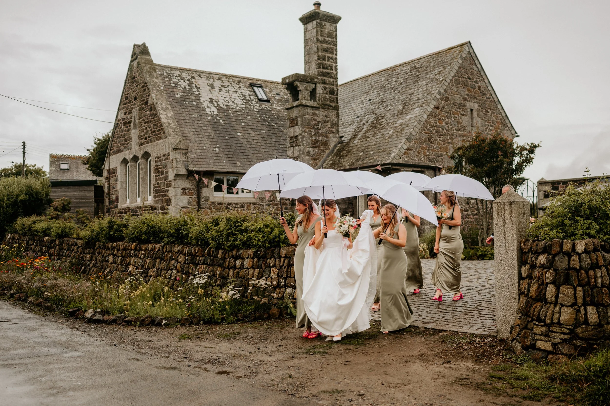 A bride and her bridesmaids walking outdoors on a cobblestone path, holding white umbrellas and flowers, in front of a historic stone building on a cloudy day.