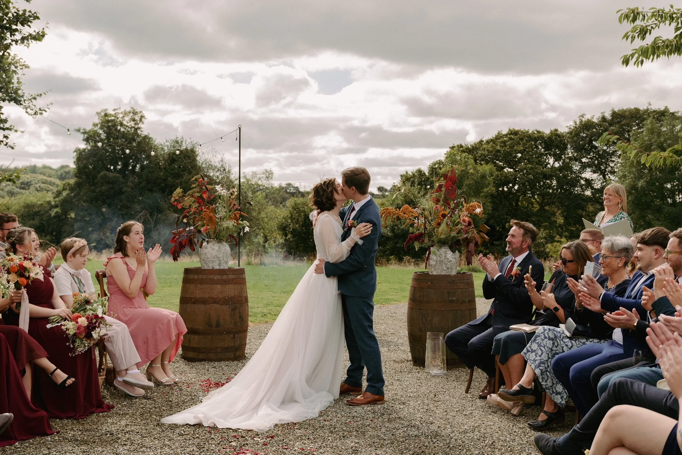 A couple gets married outdoors, kissed during their wedding ceremony, with guests clapping and smiling, surrounded by floral arrangements and trees under cloudy sky.