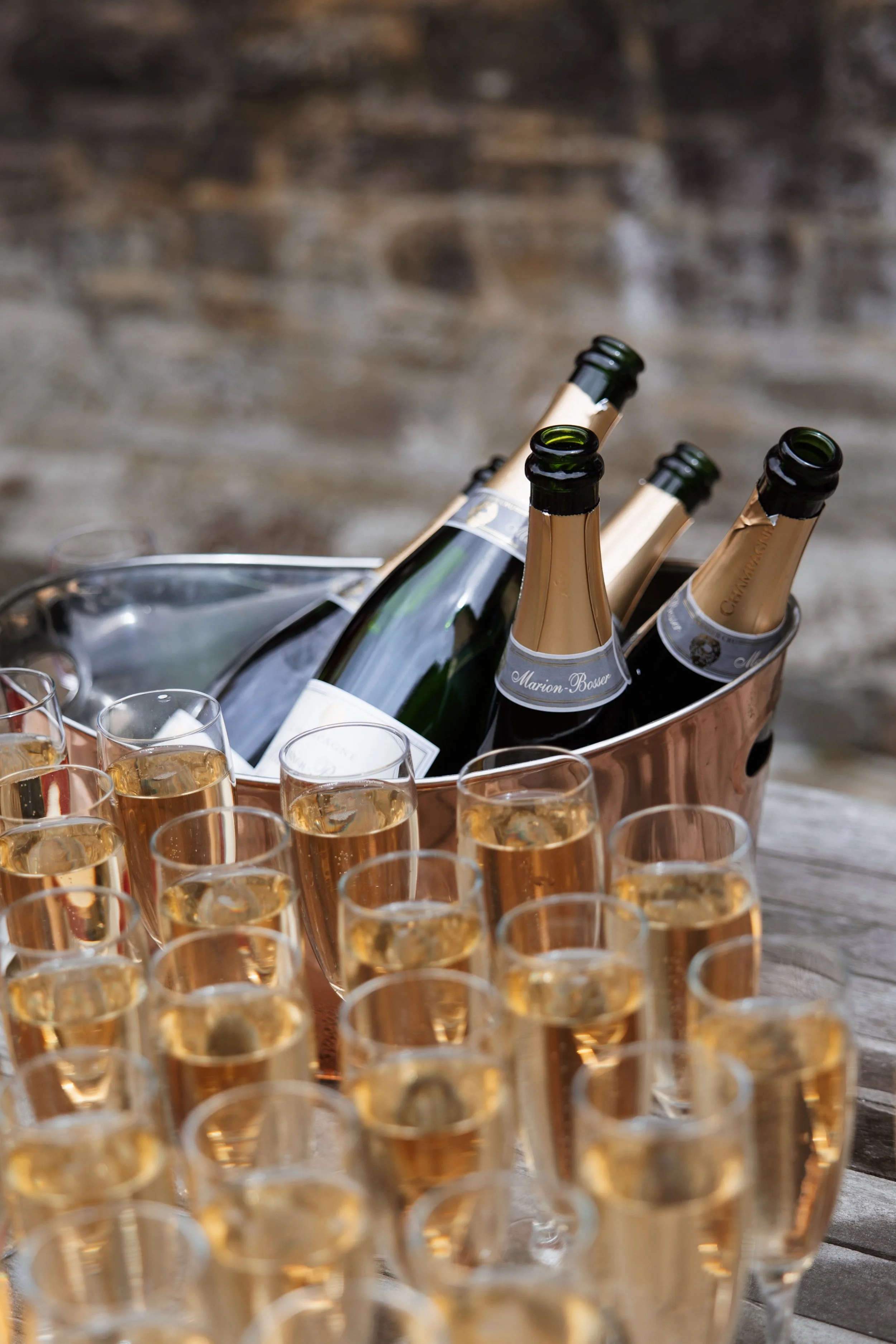 Several glasses of champagne in the foreground with a bucket of champagne bottles in the background.