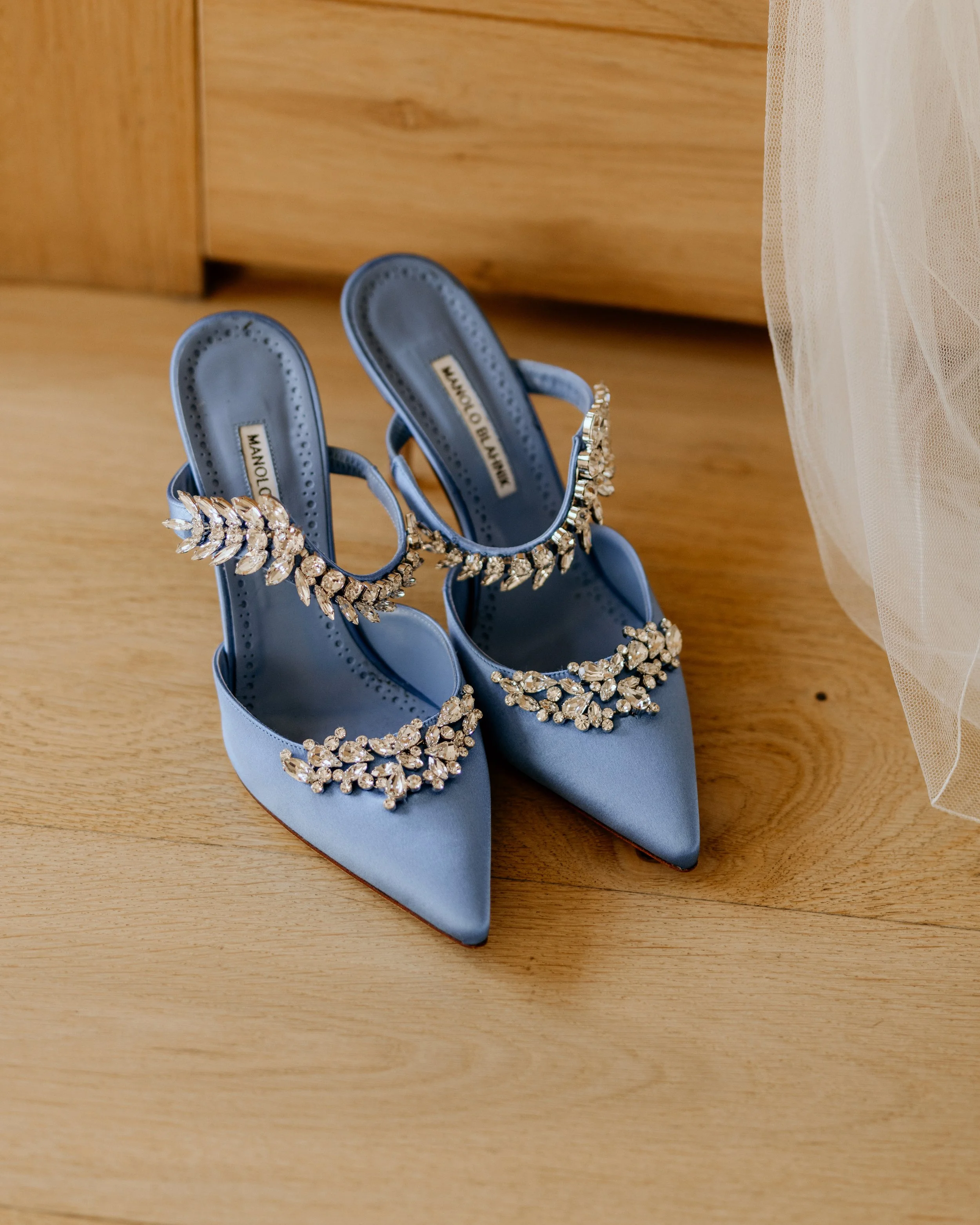 A pair of blue satin high-heeled shoes with jeweled embellishments on the straps, placed on a wooden surface next to sheer fabric.