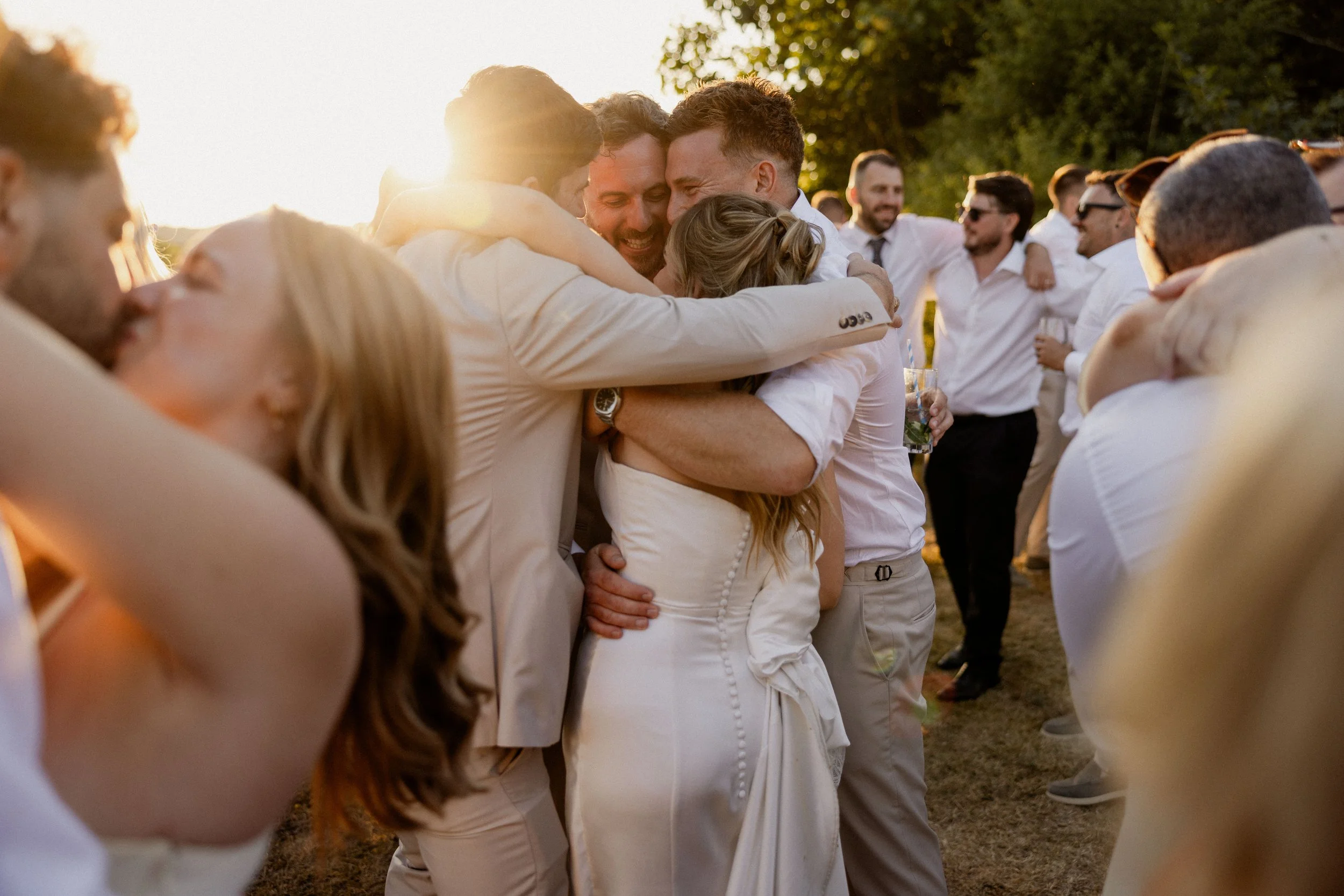 Group of people hugging and celebrating outdoors during sunset, dressed in white and light-colored clothing at a wedding or social gathering.