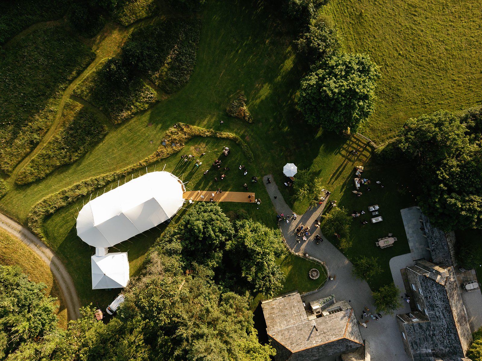 Aerial view of a garden wedding setup with a white tent, a wooden aisle, and guests gathered on the grass.