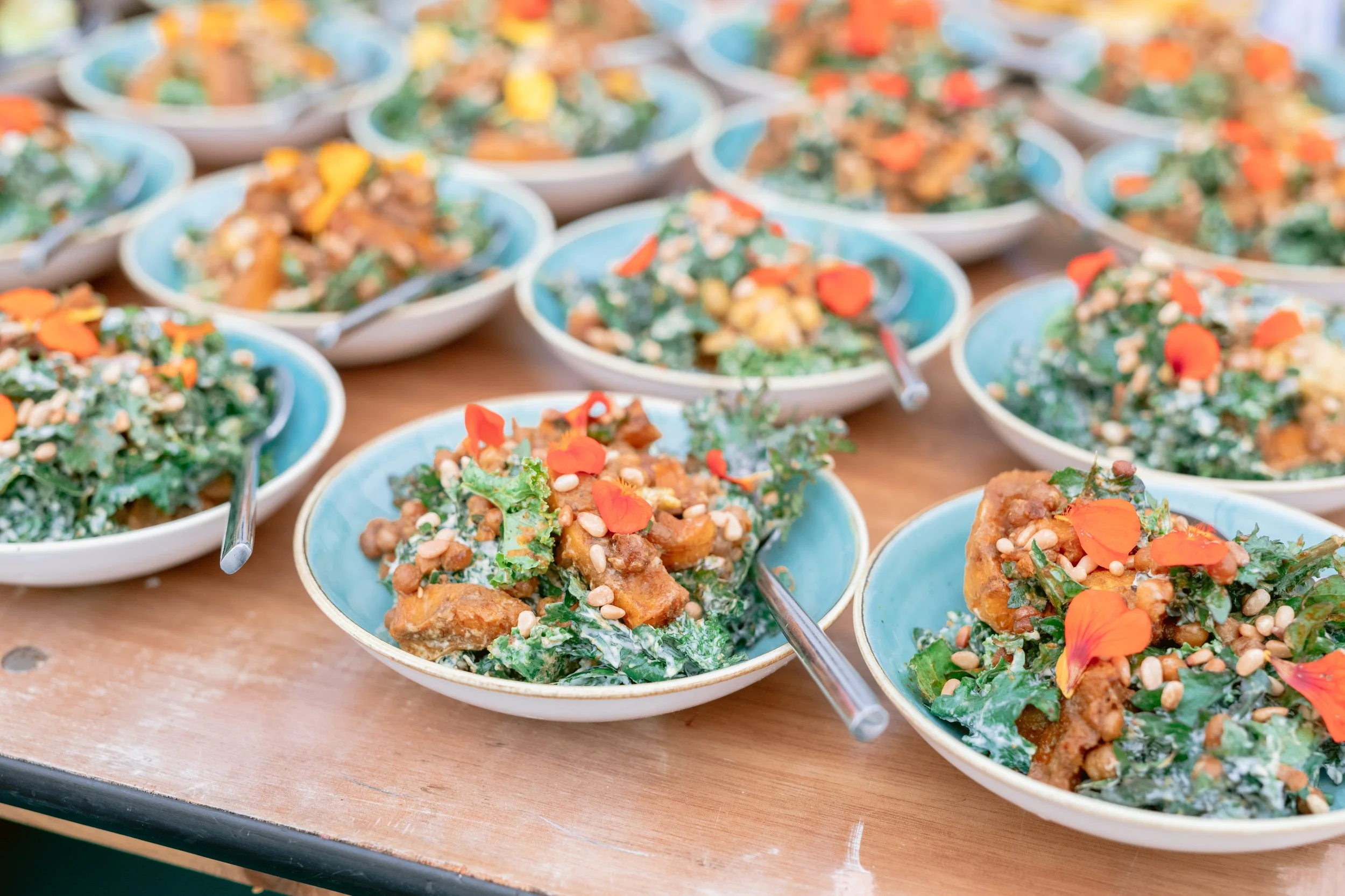 Several bowls of a salad with leafy greens, roasted croutons, sunflower seeds, and orange flower petals on top, placed on a wooden table, with metal serving spoons.