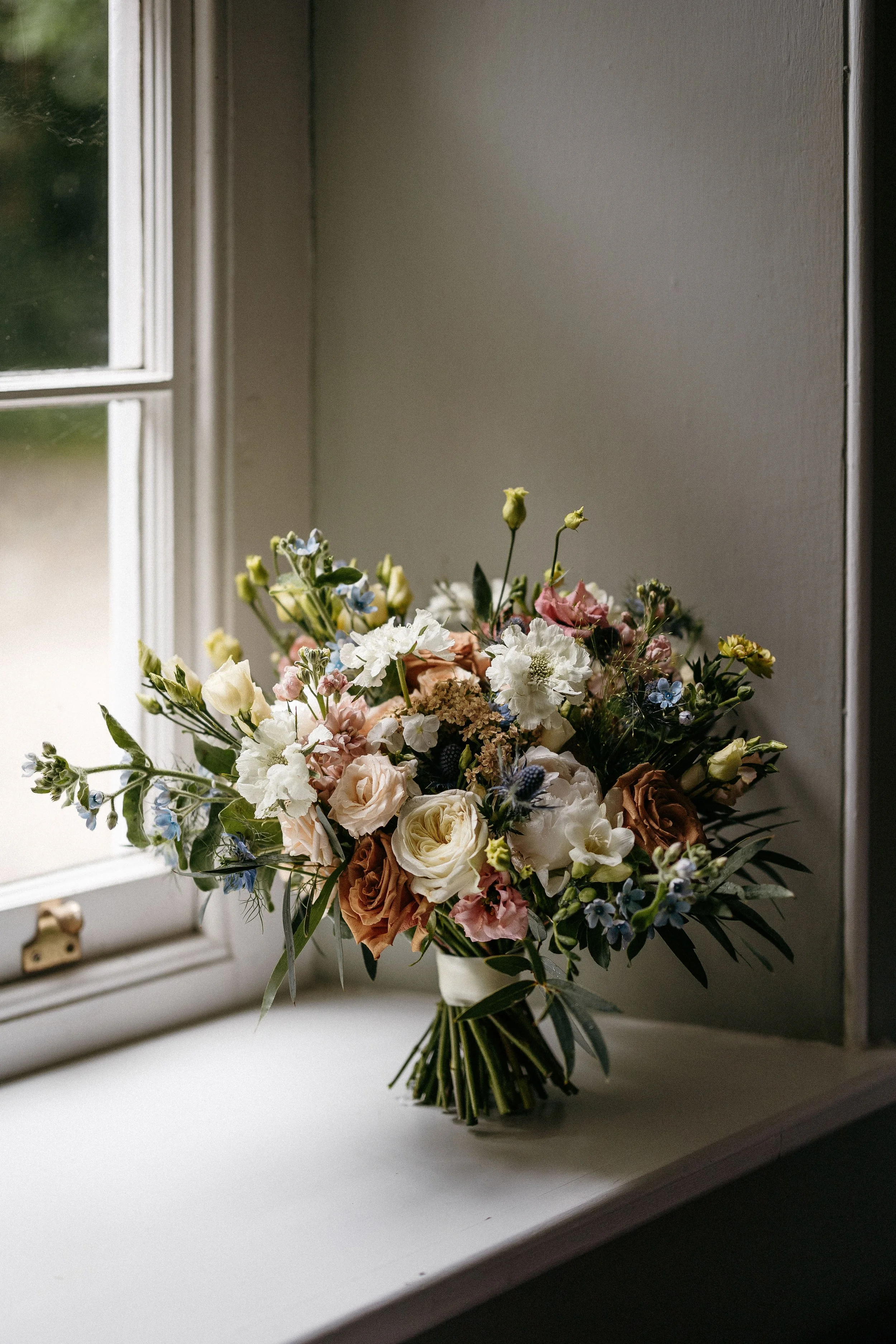 A bouquet of mixed flowers, including roses, on a windowsill near a window with natural light.