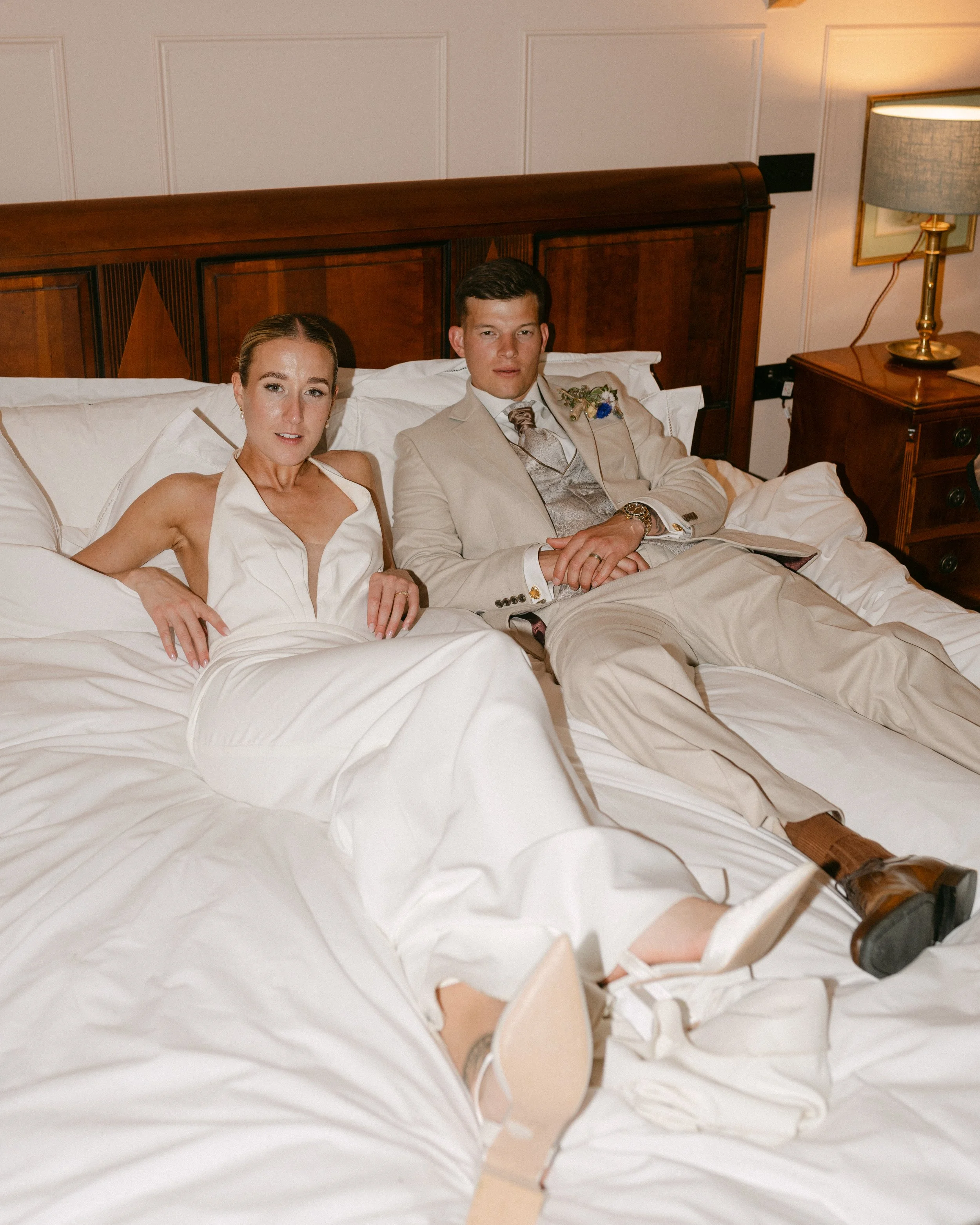 A woman and a man lying on a bed in wedding attire in a warmly lit room with wooden furniture and white paneled walls.