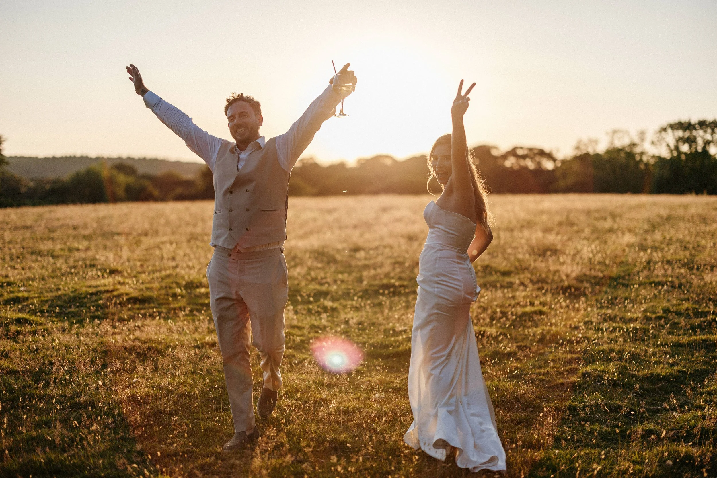 A couple in wedding attire celebrating outdoors at sunset, holding drinks and making peace signs.