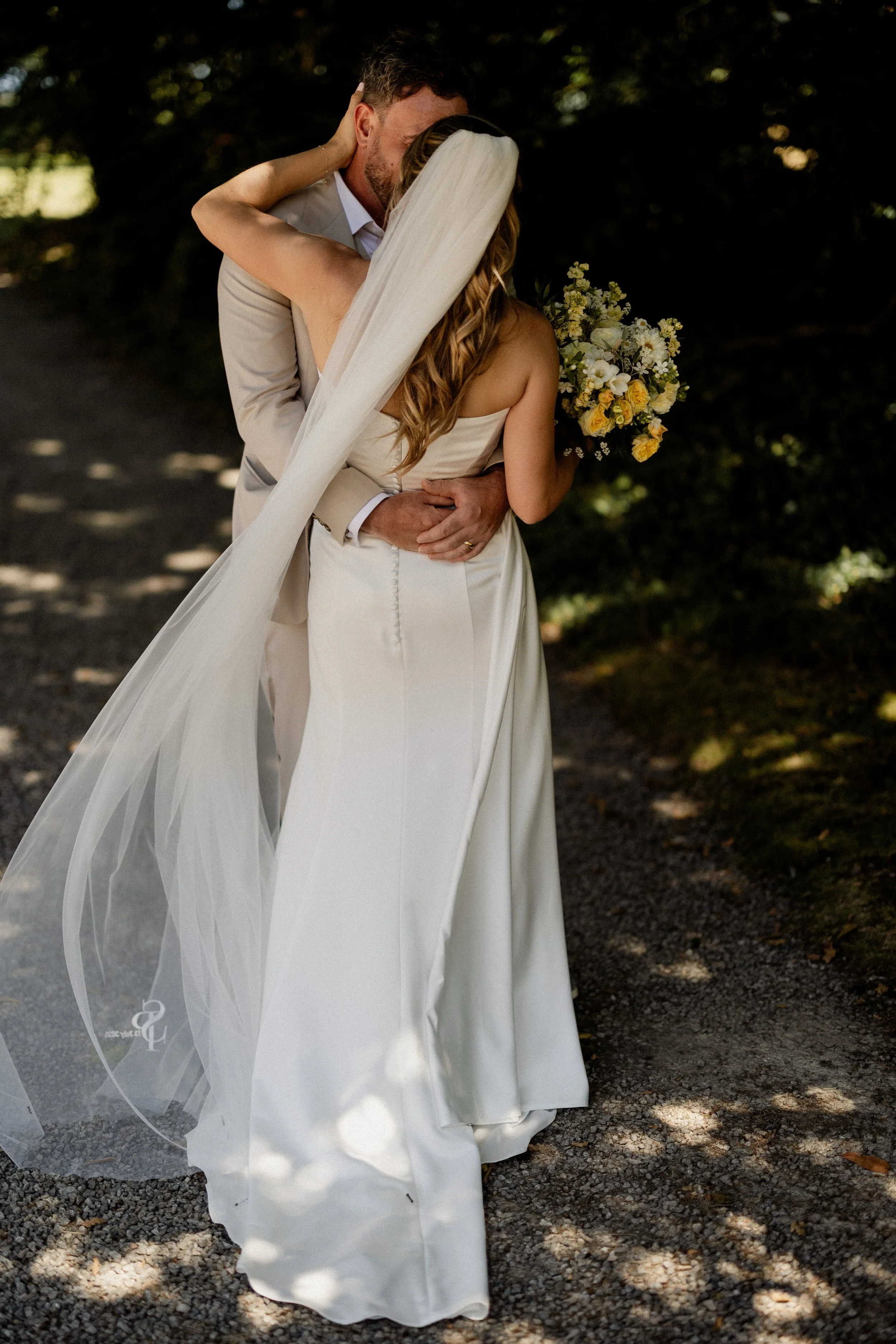 A bride and groom share an intimate moment outdoors, with the groom kissing the bride's forehead. The bride is holding a bouquet of white and yellow flowers, and she has long, wavy hair and a veil. The groom is wearing a light-colored suit, and they are standing on a shaded gravel pathway surrounded by trees.