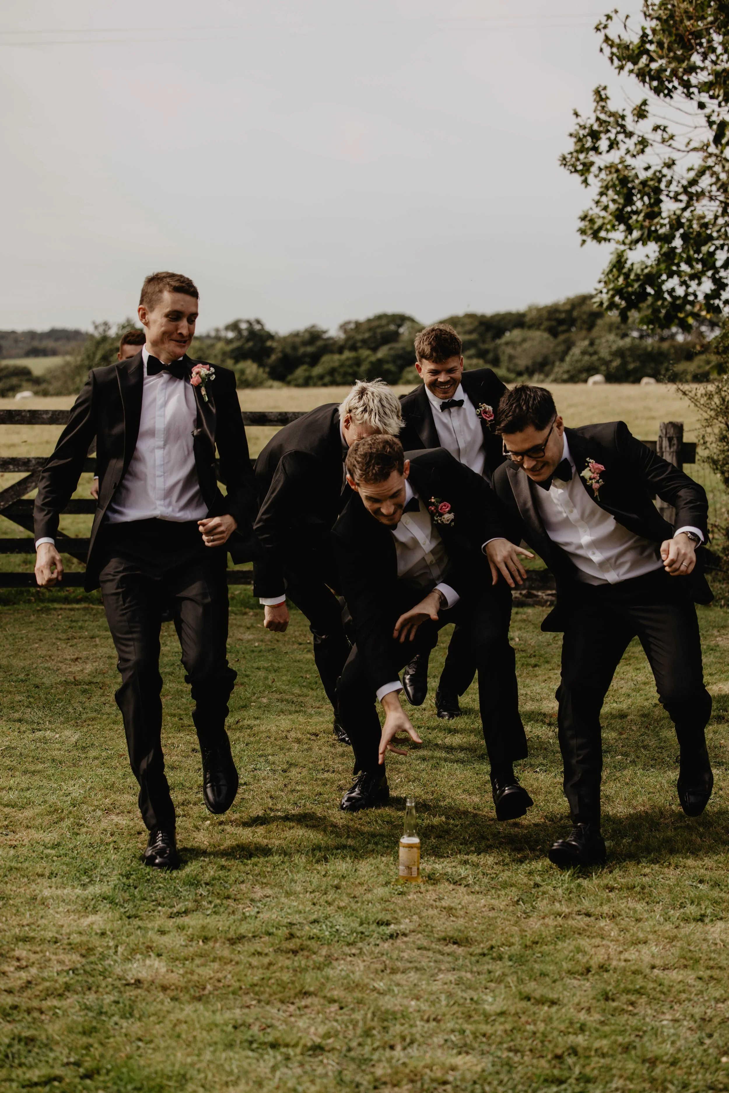 Group of five men in tuxedos playing a game outside on grass, with one bottle on the ground, in a rural setting with trees and a fence in the background.