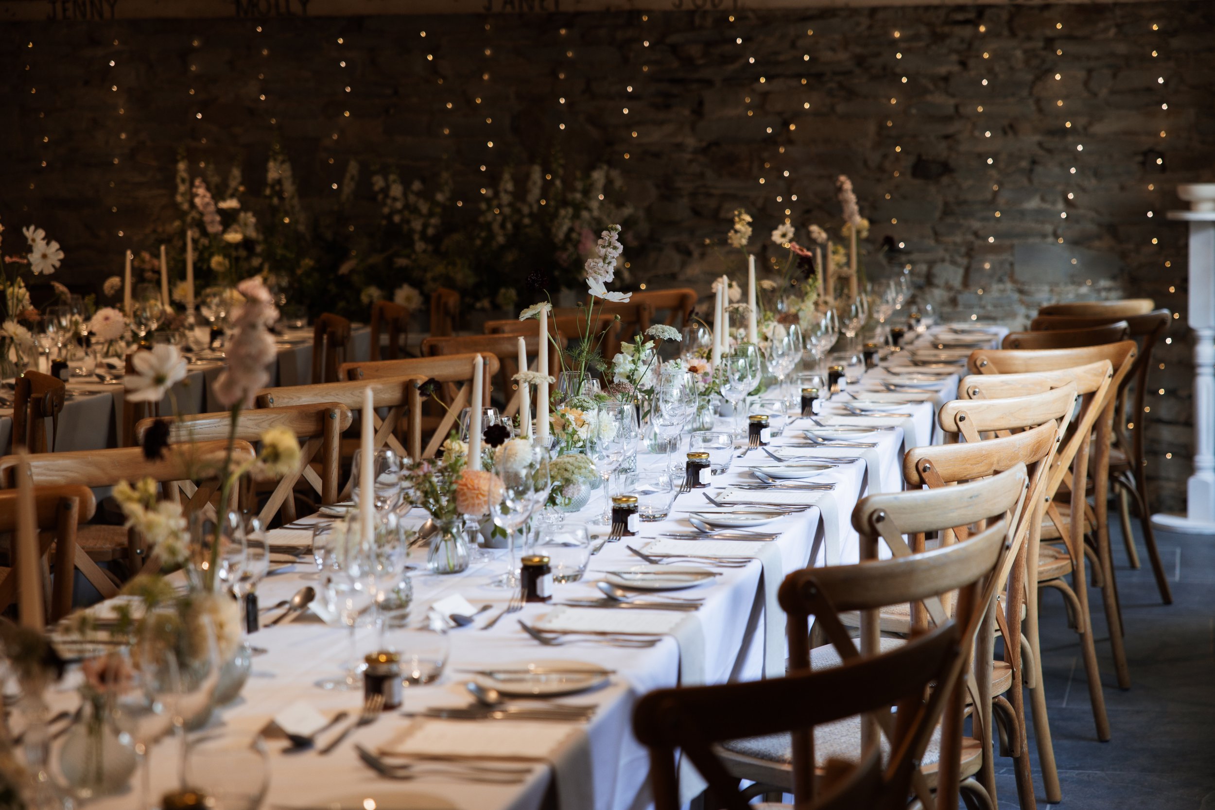 Long dining table set for a wedding or special event with white tablecloths, floral centerpieces, candles, and glassware, against a stone wall decorated with fairy lights.