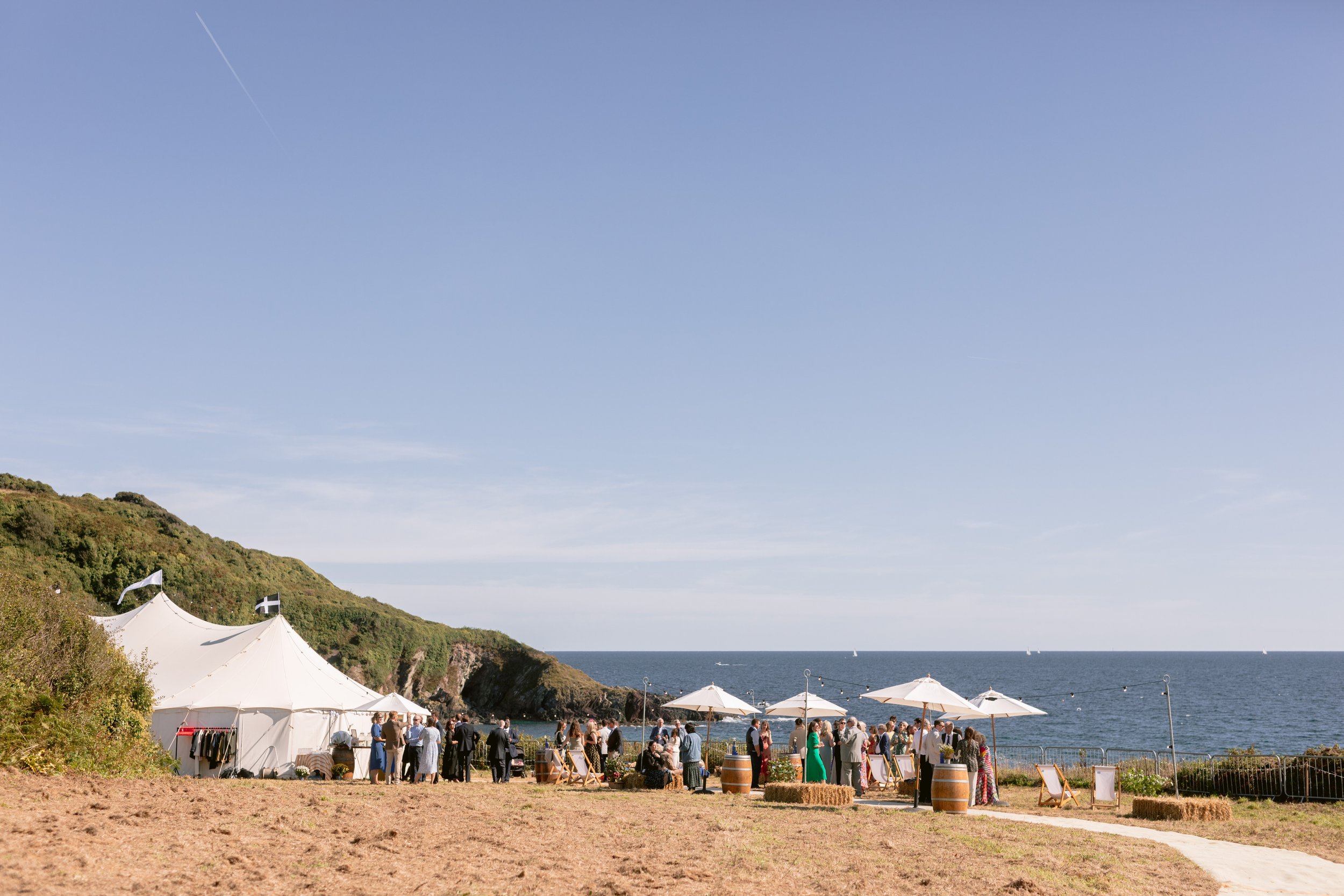 A beachside outdoor event with a large white marquee tent, smaller umbrellas, and groups of people gathered on a sandy area with a grassy hillside and ocean in the background on a sunny day.