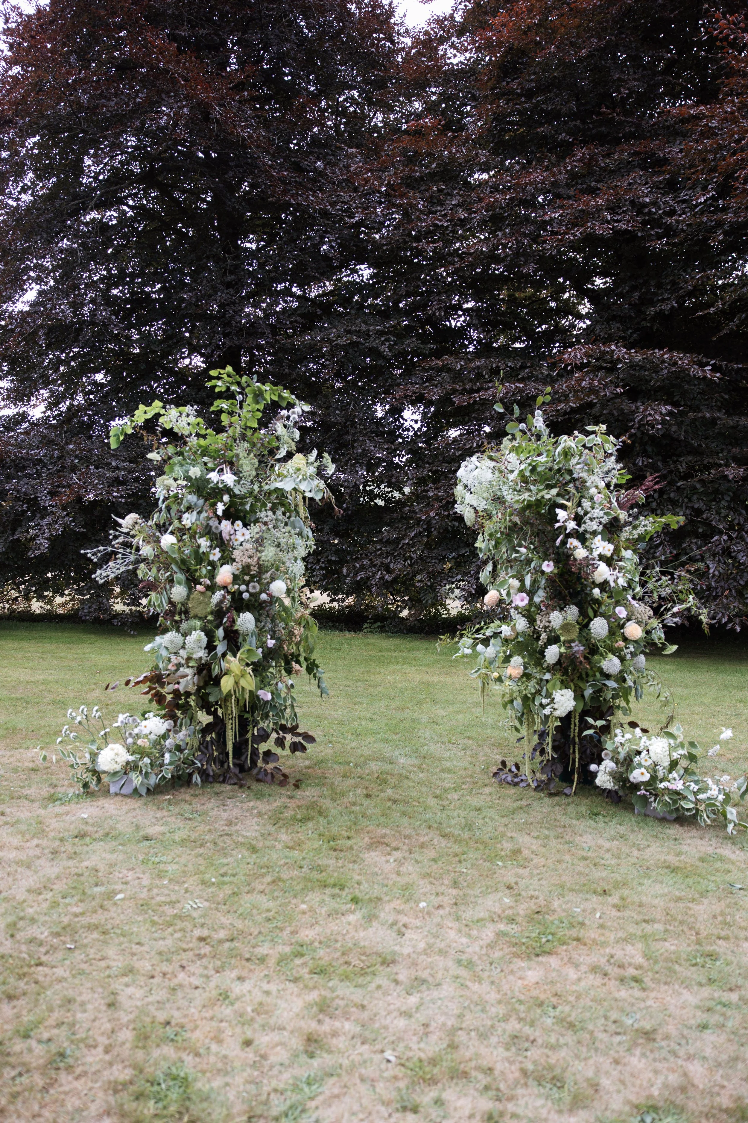 Two large floral arrangements with white flowers and greenery, set outdoors on a grassy area with dark trees in the background.