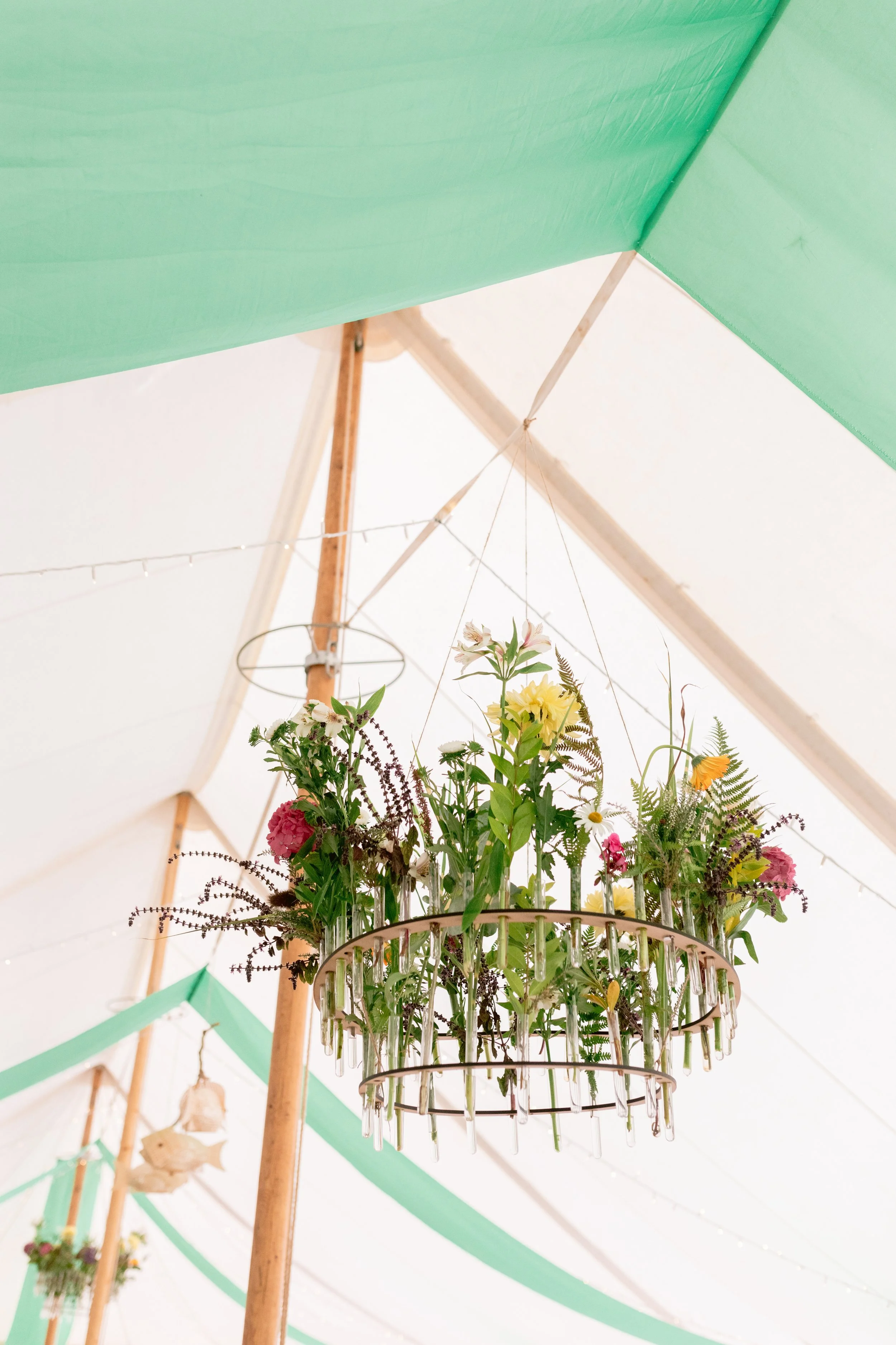 Chandelier made with flowers and glass tubes hanging from a circular metal frame, decorated with green leaves and various colorful flowers, hanging under a tent with green fabric and string lights.