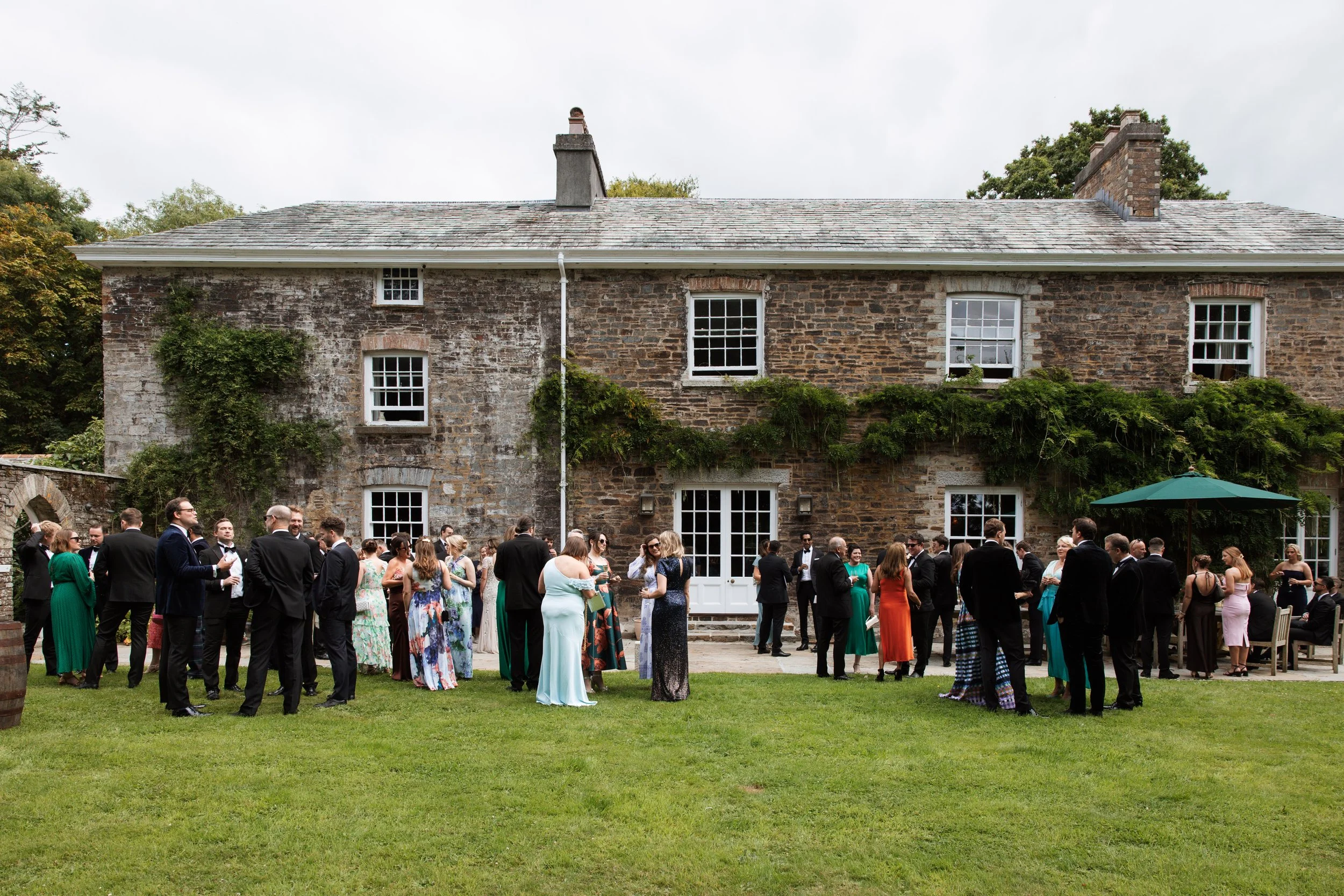 Guests in formal attire gather outside near a rustic stone house during a celebration or wedding reception.